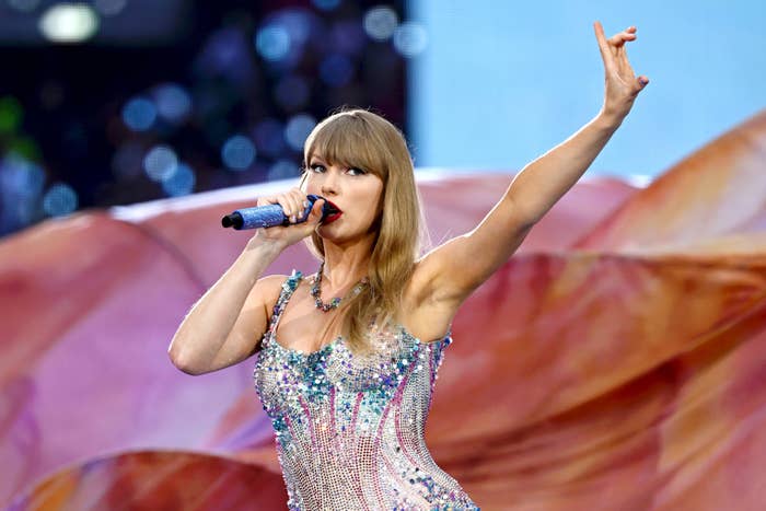 Taylor Swift performs on stage, holding a microphone with a confident pose. She wears a sparkling, fitted dress