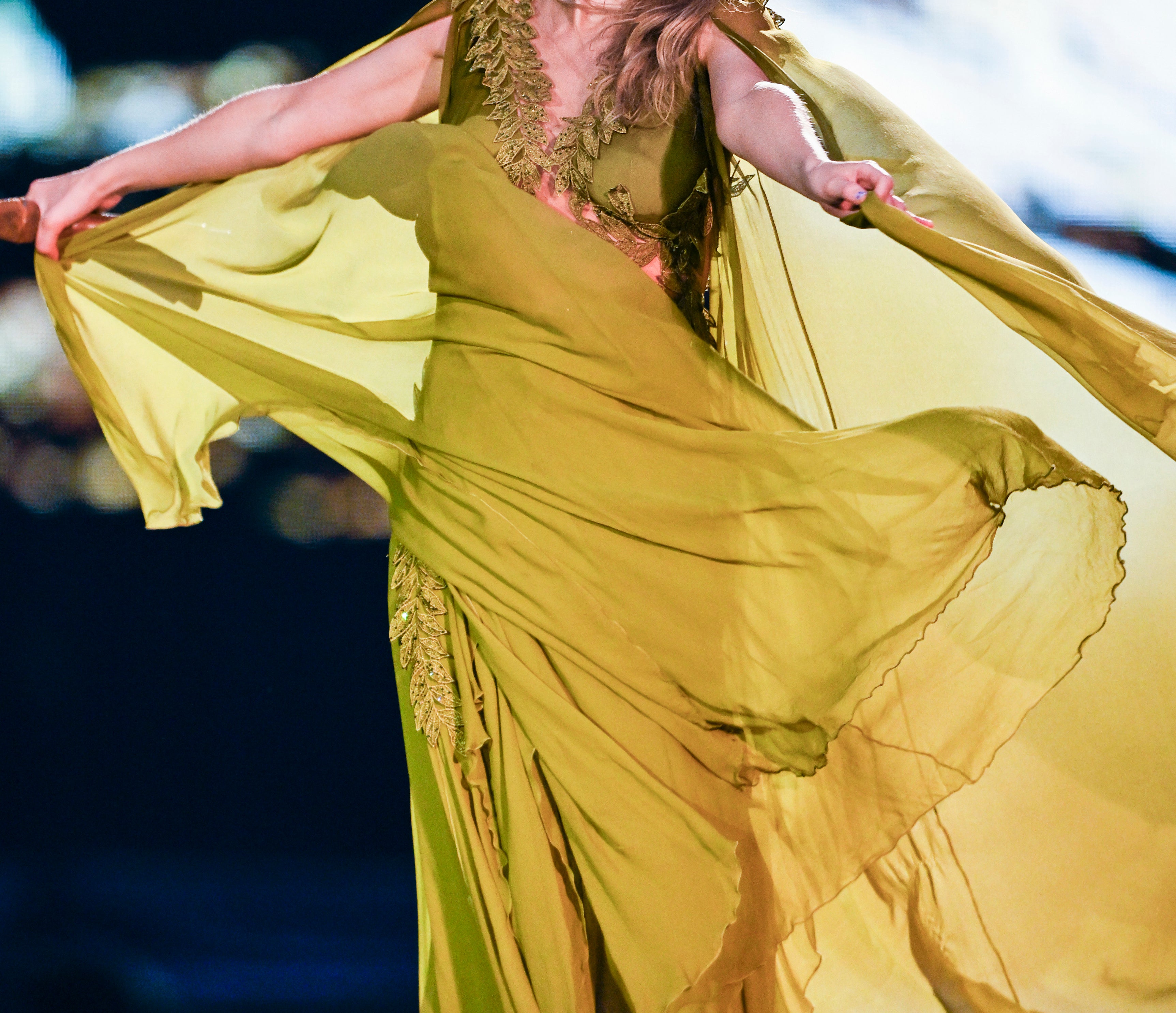 Taylor Swift performing on stage, wearing a flowing, elegant dress with intricate patterns