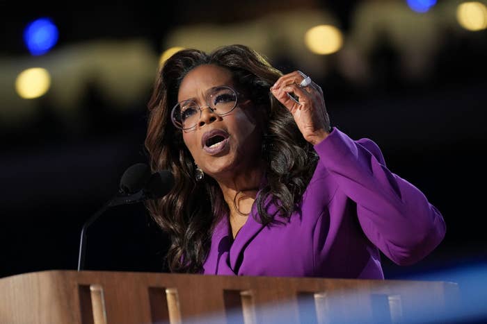 Oprah Winfrey speaks passionately from a podium. She wears a stylish outfit and glasses, gesturing with one hand raised