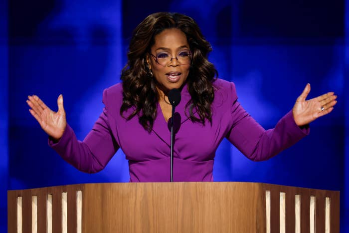 Oprah Winfrey stands at a podium, gesturing with both hands while speaking. She is wearing a stylish, fitted suit