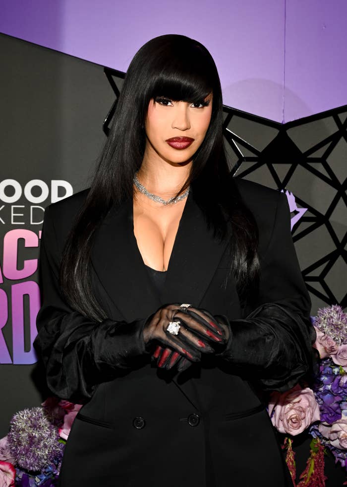 Cardi B poses on the red carpet in a sleek dark outfit with gloves, a plunging neckline, and straight hair with bangs at an event