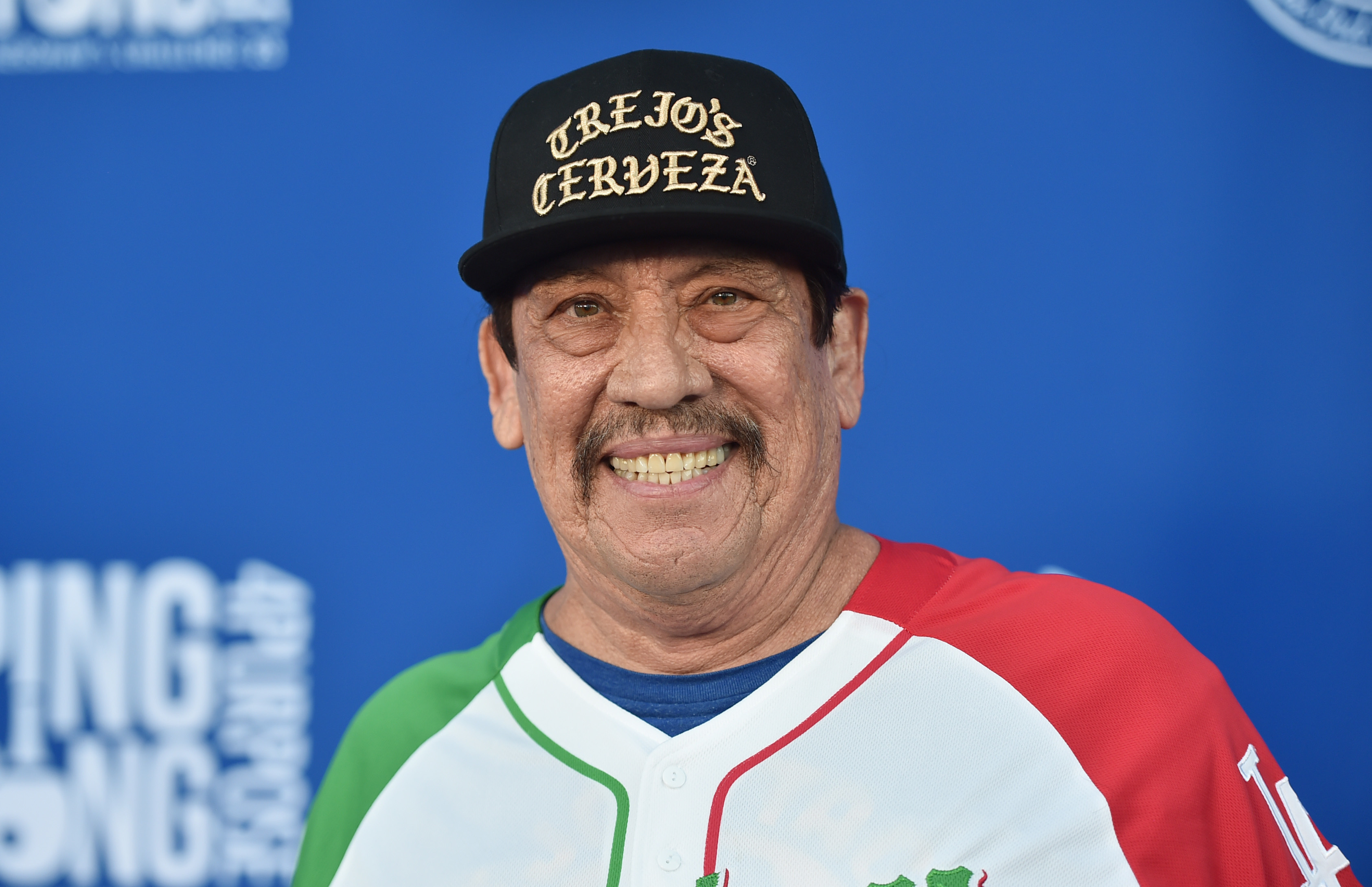 Danny Trejo smiles while wearing a "Trejo's Cerveza" cap and a baseball jersey