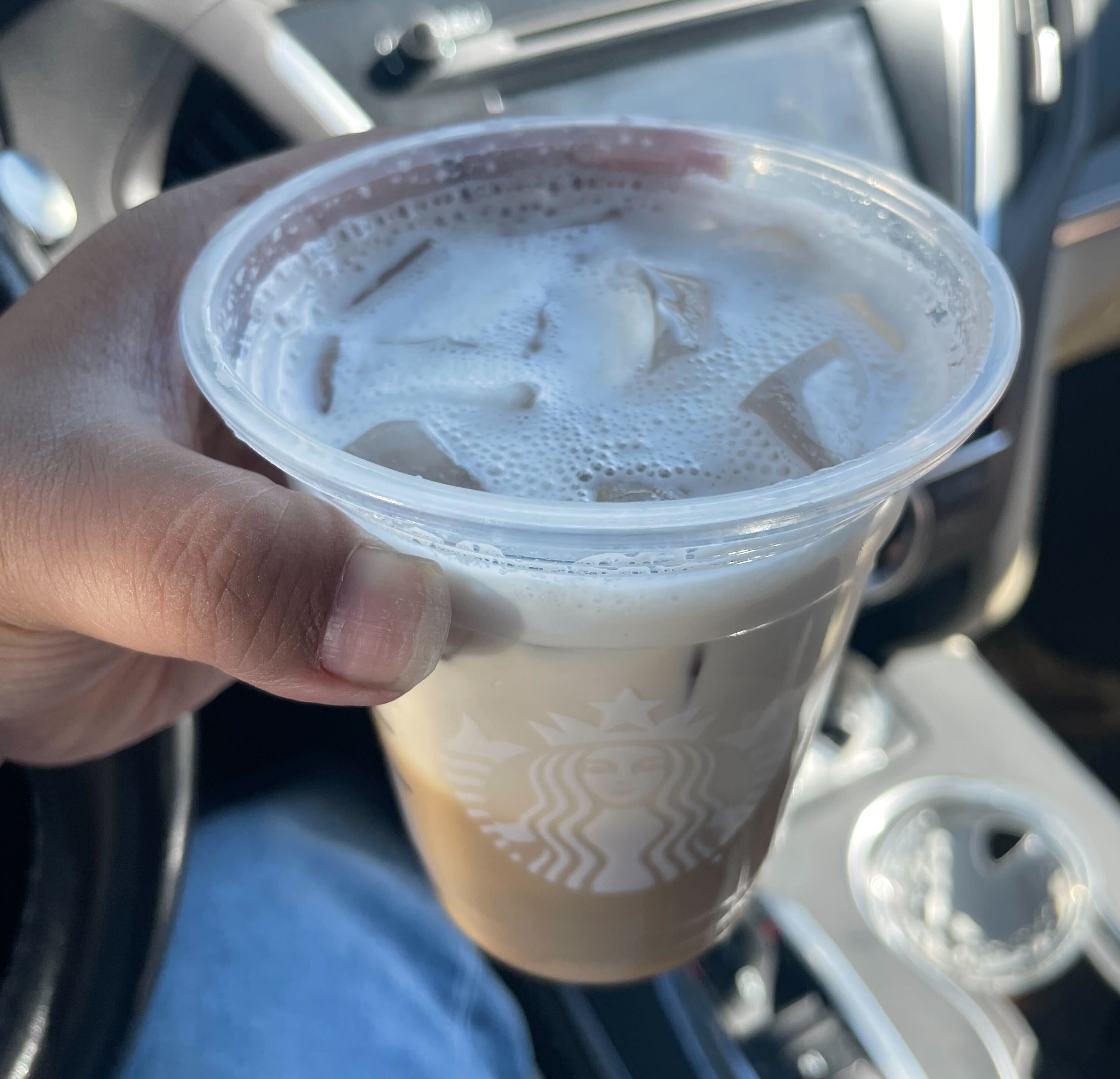 A person's hand holding an iced coffee beverage with foam in a clear plastic cup inside a car