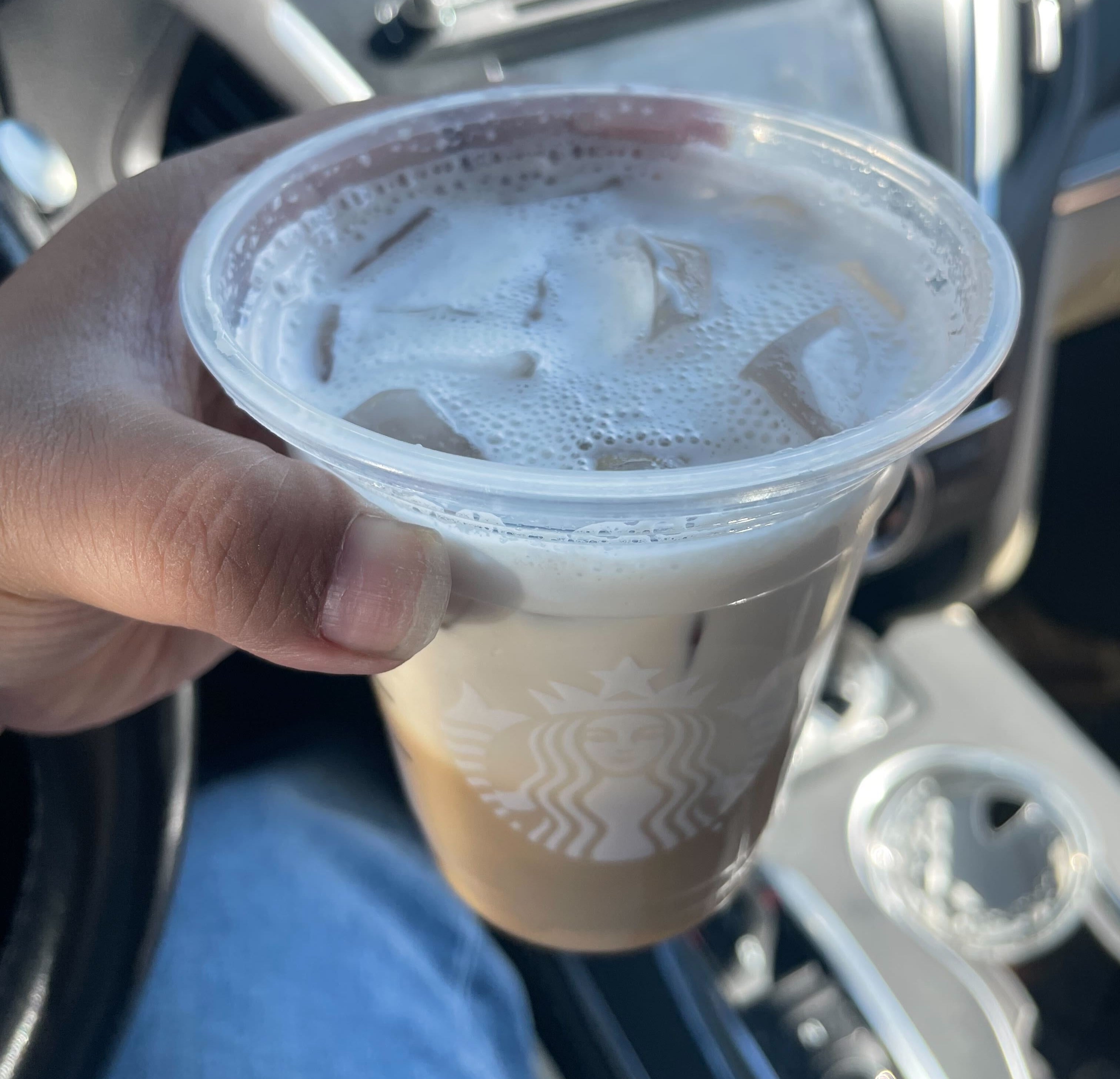A person's hand holding an iced coffee beverage with foam in a clear plastic cup inside a car