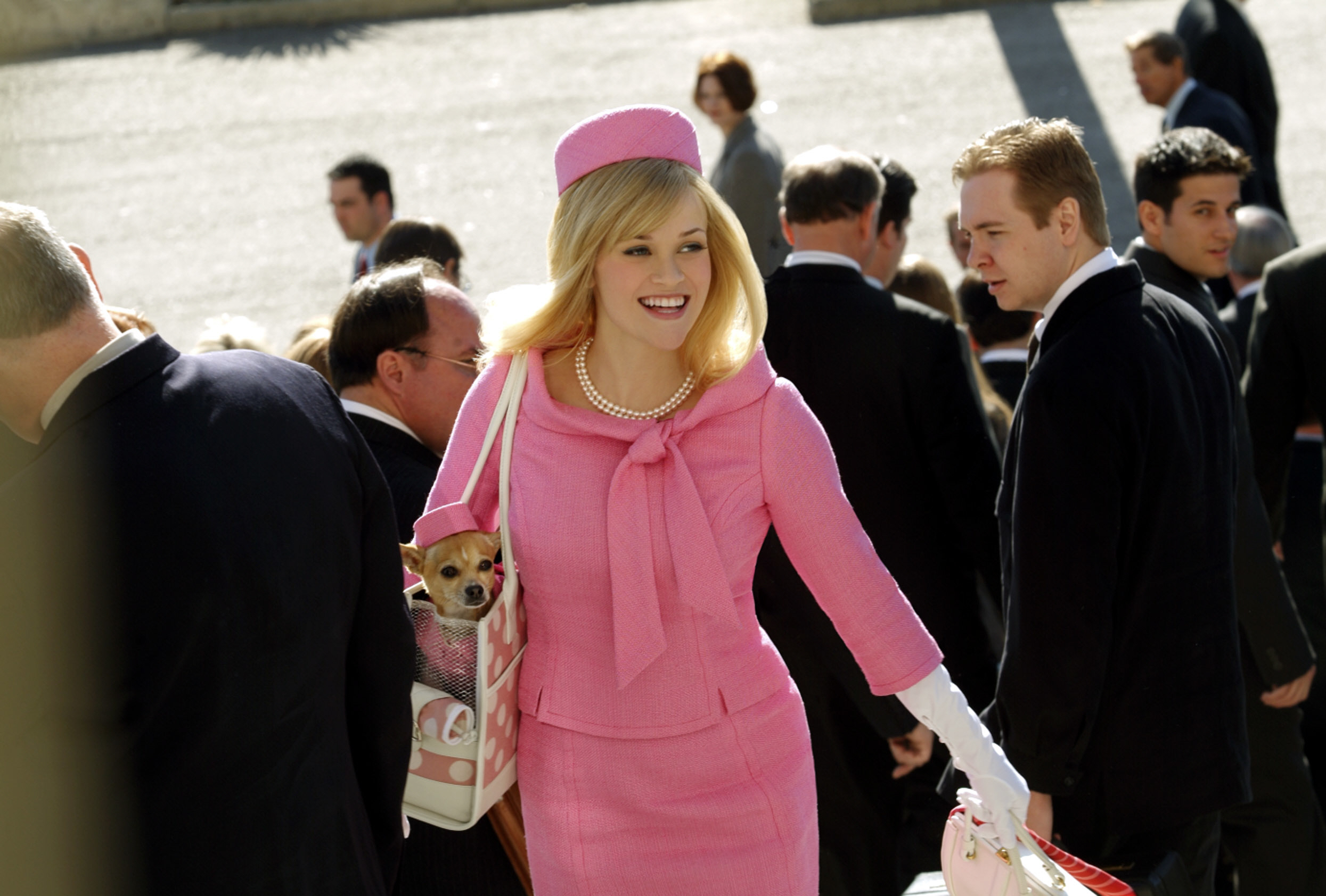 Reese Witherspoon, dressed in a stylish suit and matching hat, smiles while holding a small dog in her bag among a crowd