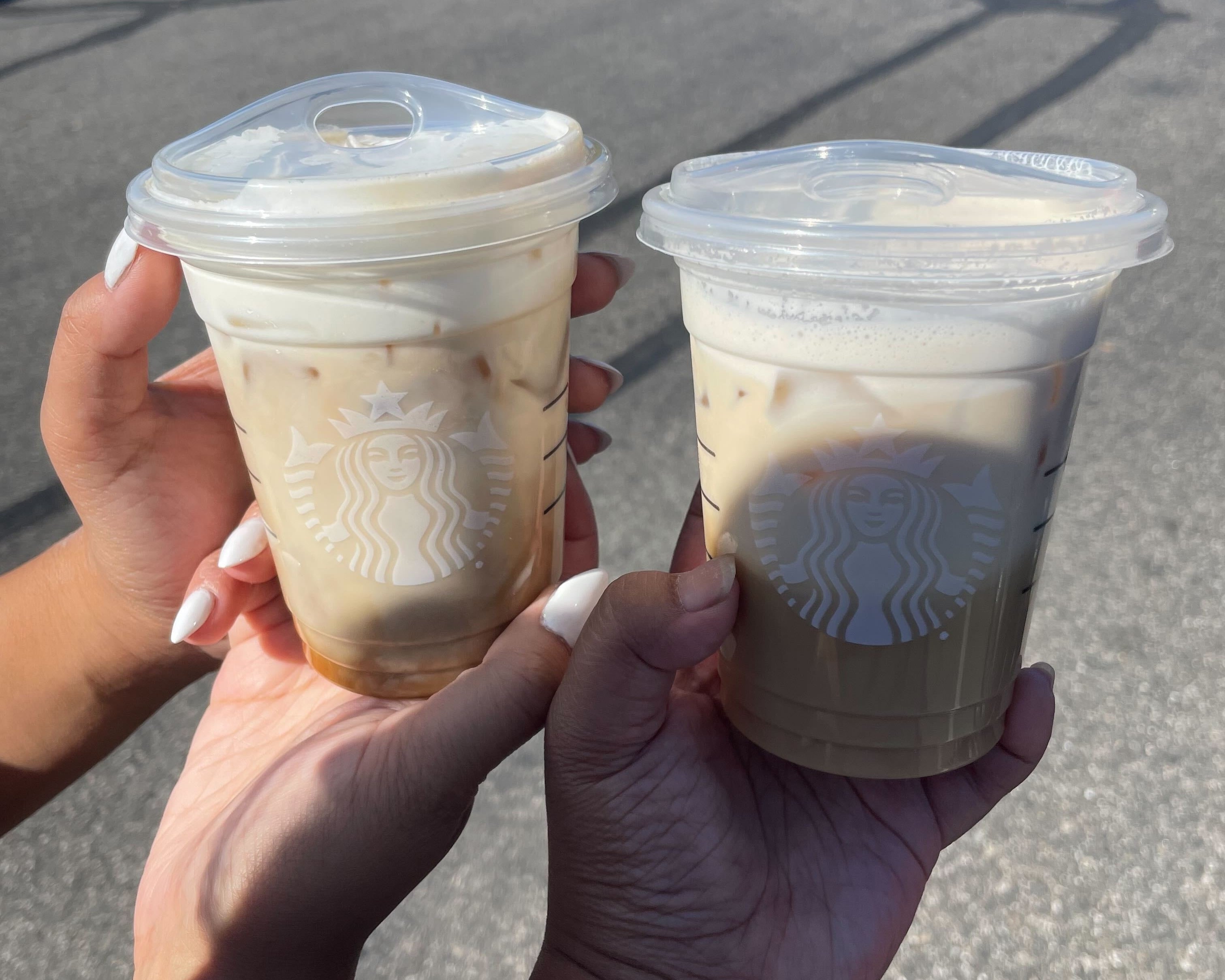 Two hands holding Starbucks iced coffee drinks with clear plastic lids in a parking lot on a sunny day