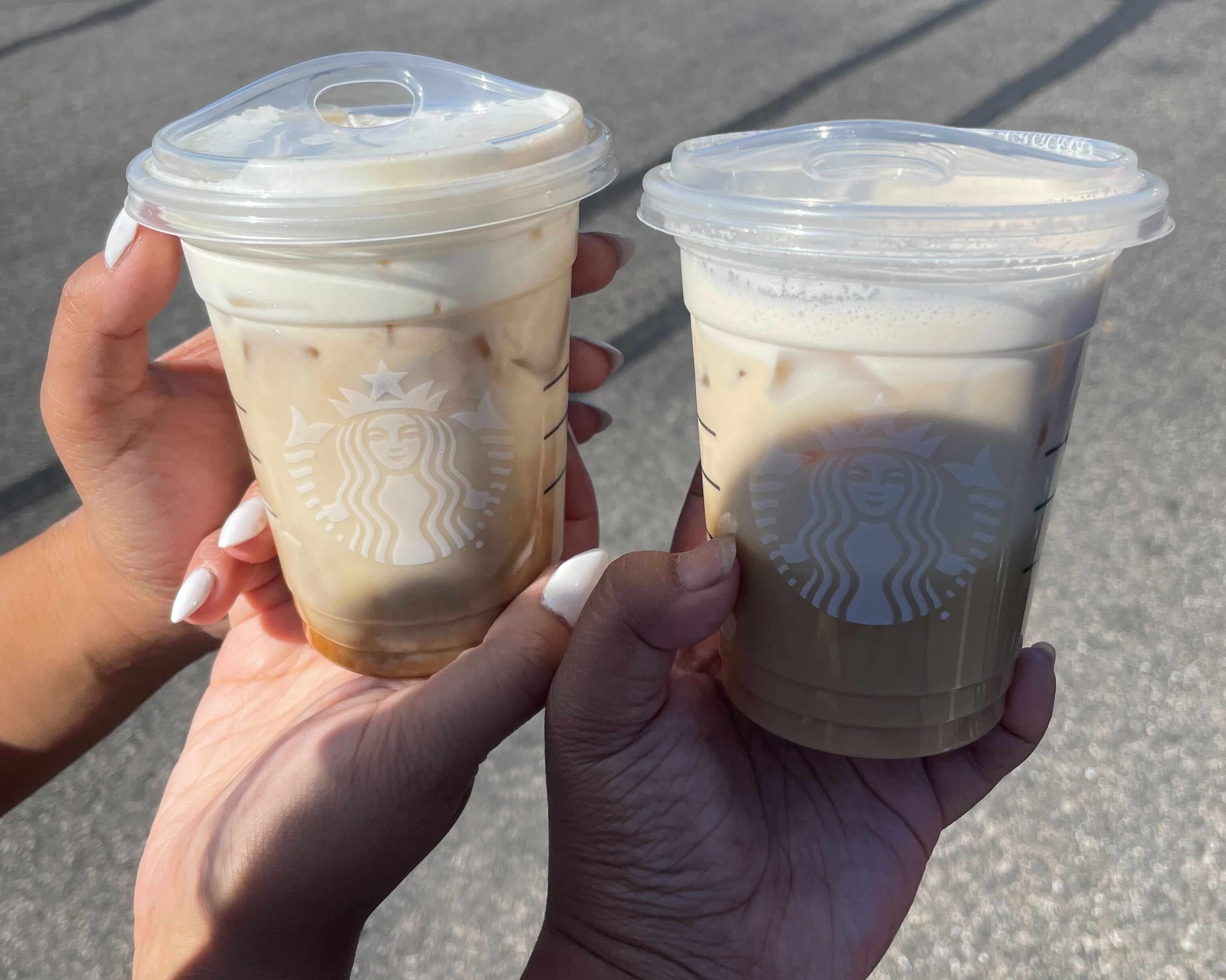 Two hands holding Starbucks iced coffee drinks with clear plastic lids in a parking lot on a sunny day