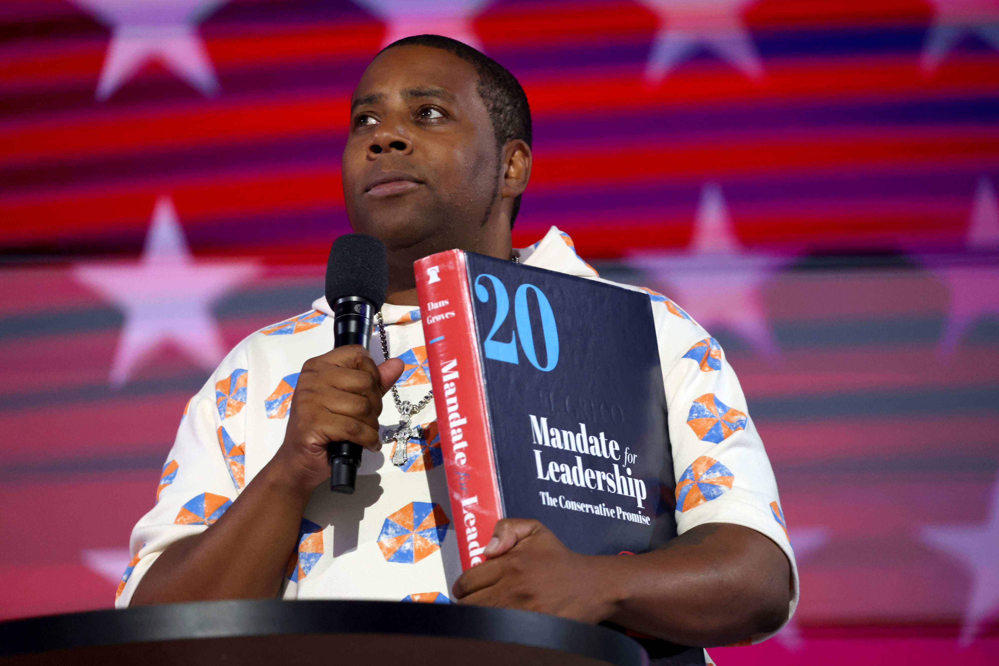 Kenan Thompson holds a microphone and a large book titled "Mandate for Leadership" at an event. Stars and stripes pattern backdrop in the background