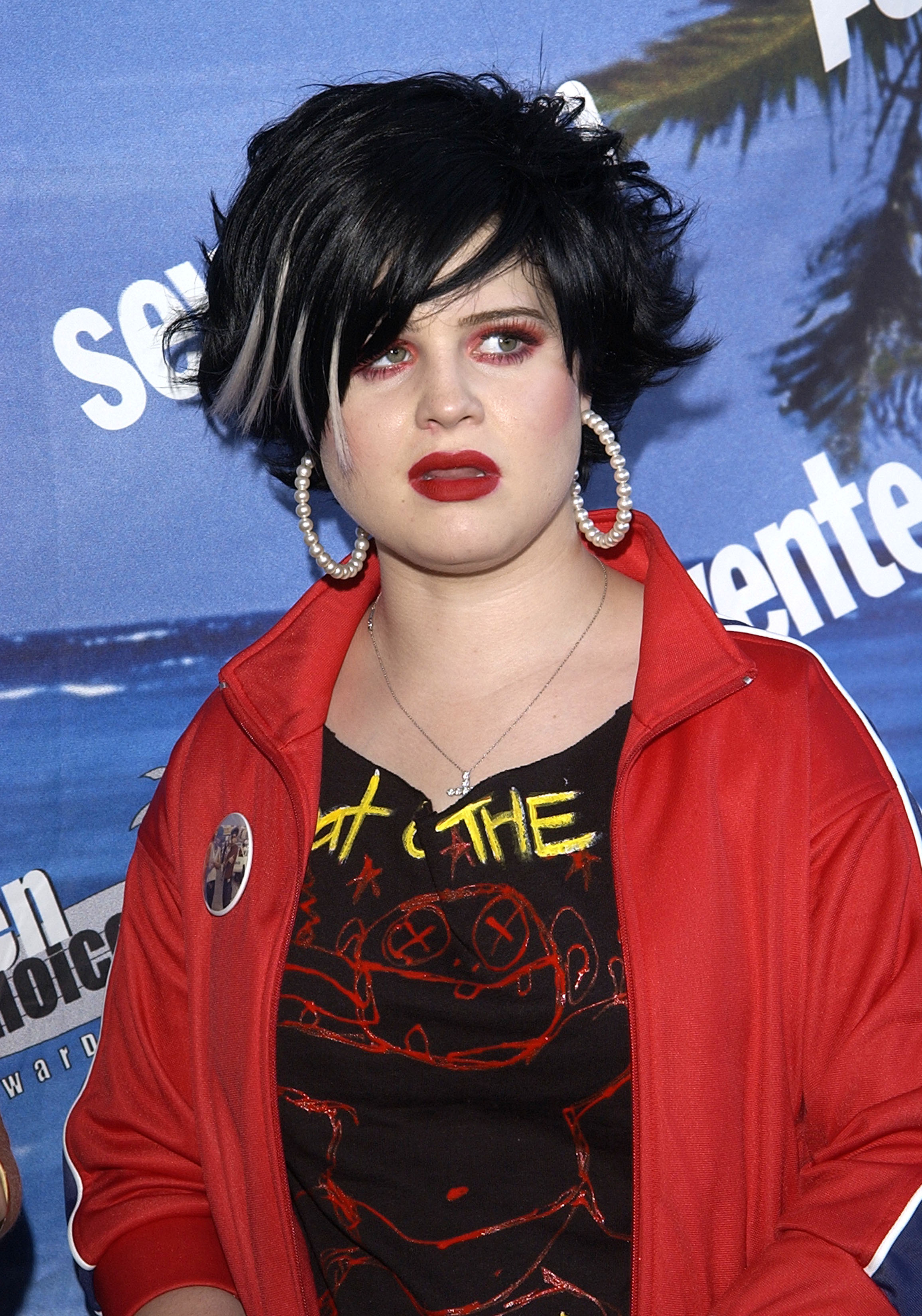 Kelly Osbourne in punk-inspired makeup with a voluminous bob haircut, wearing a jacket with stripes and printed T-shirt, accessorized with large hoop earrings