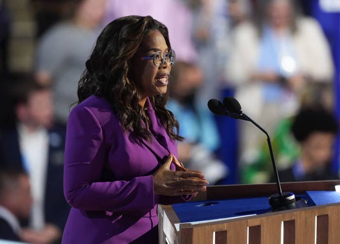Oprah Winfrey speaking at a podium, wearing a stylish blazer and glasses, addressing an audience at an event