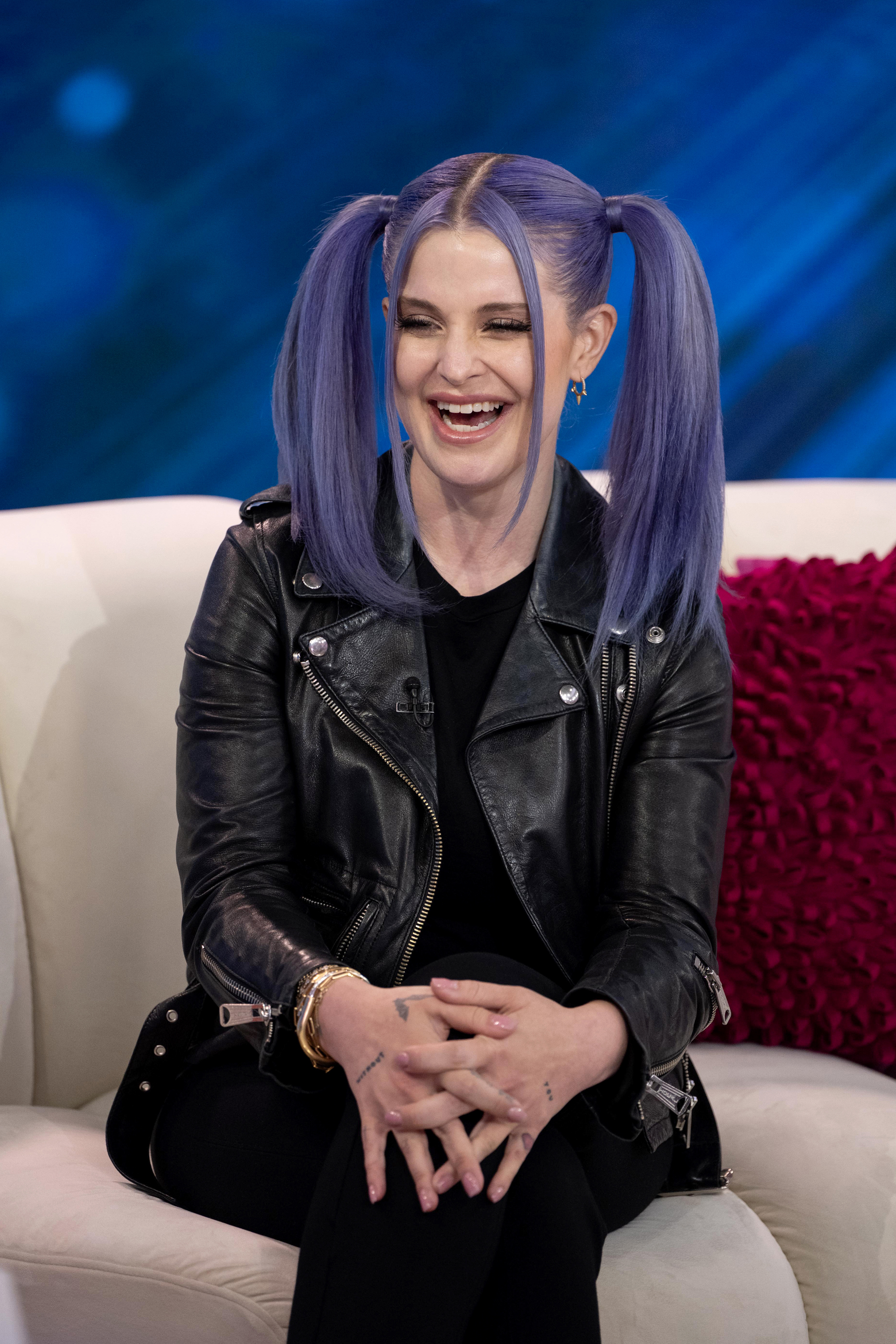 Kelly Osbourne is seated on a couch, smiling widely. She has her hair styled in two high pigtails and is wearing a black leather jacket