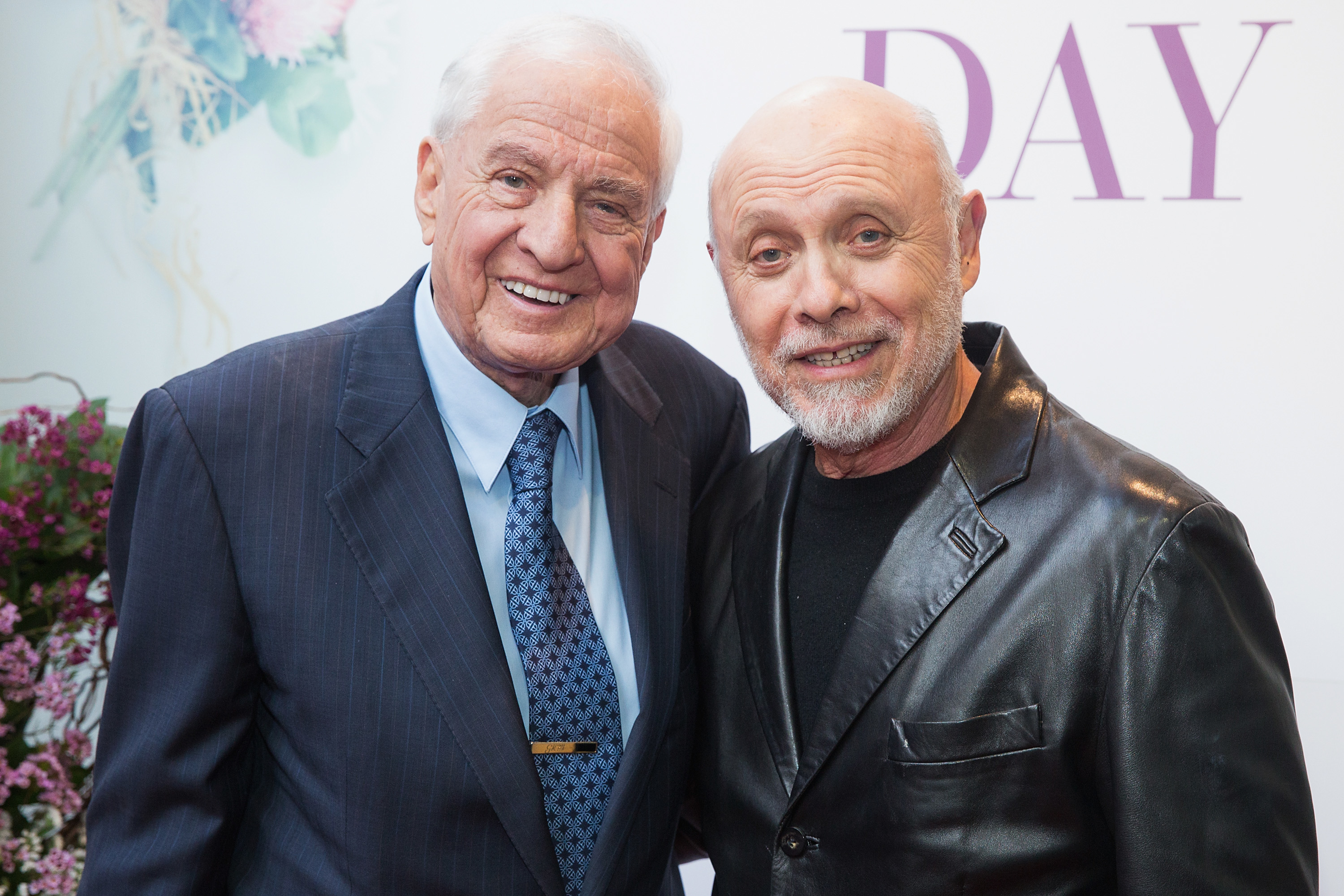 Garry Marshall in a suit and tie stands next to Héctor Elizondo wearing a leather jacket, both smiling at the camera