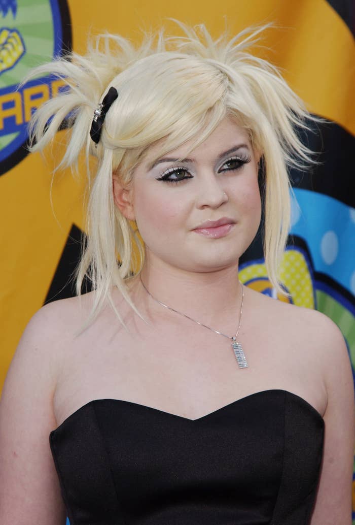 Kelly Osbourne poses on a red carpet, wearing a strapless dress with unique spiked hair styled with a small black bow