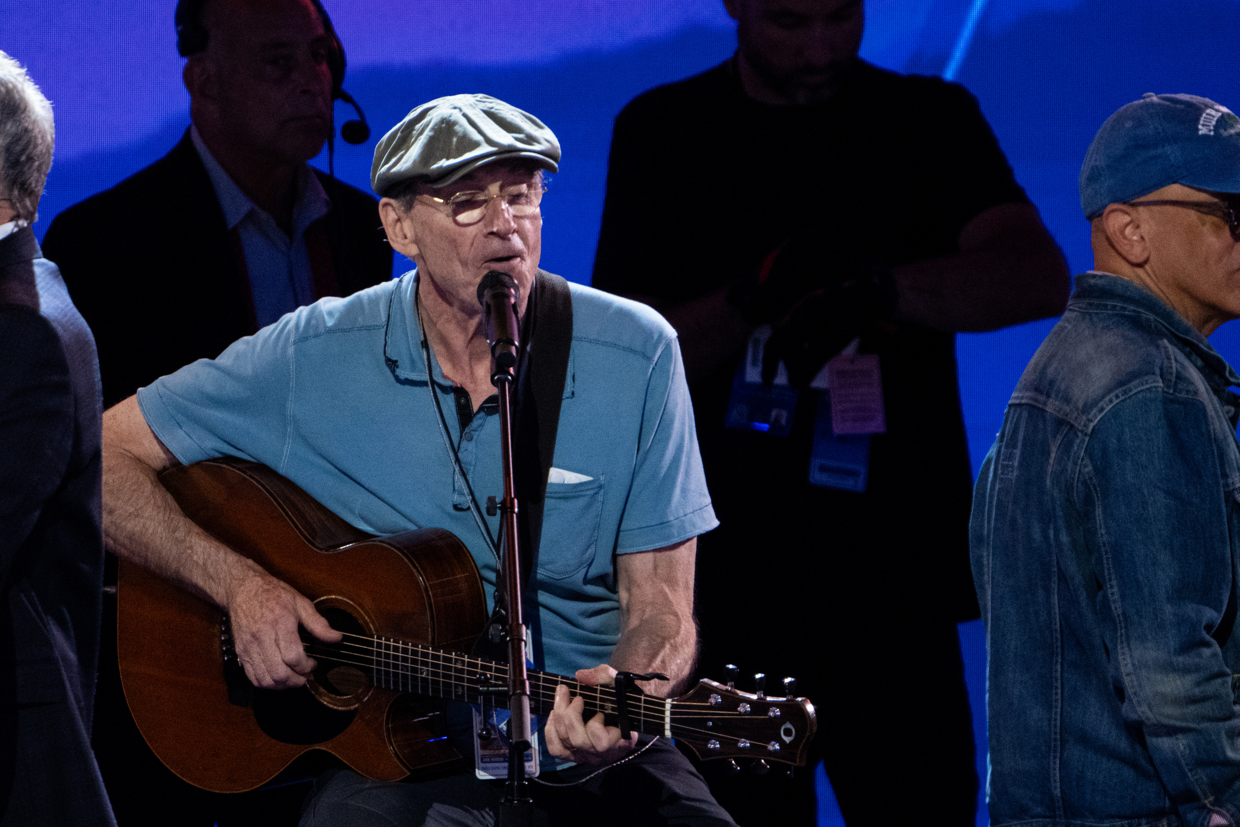 James Taylor performs on stage, wearing a cap and casual attire, playing an acoustic guitar. Several people are visible in the background