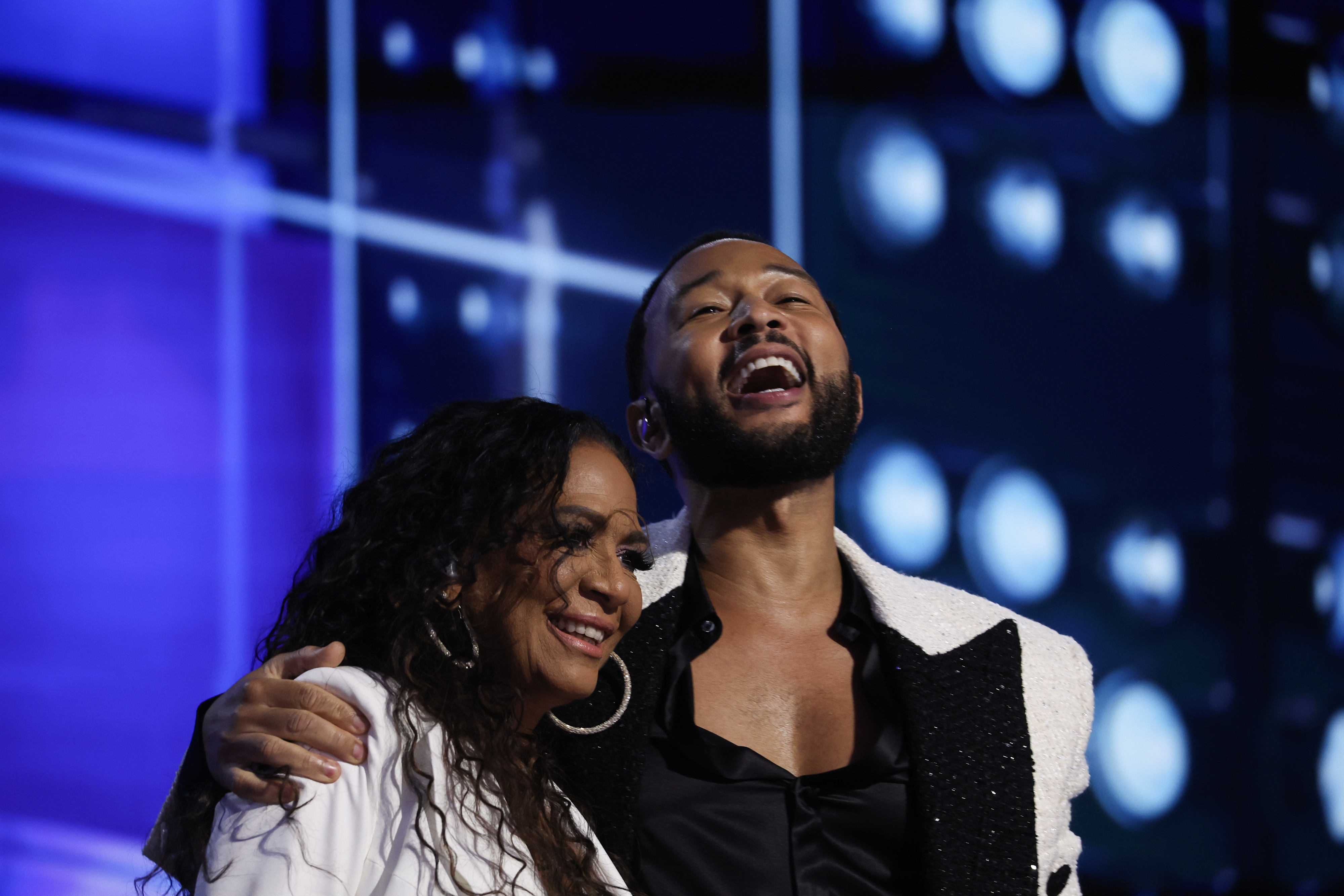 John Legend and Joy Enriquez sharing a happy moment on stage, John in a stylish white jacket with a black lapel