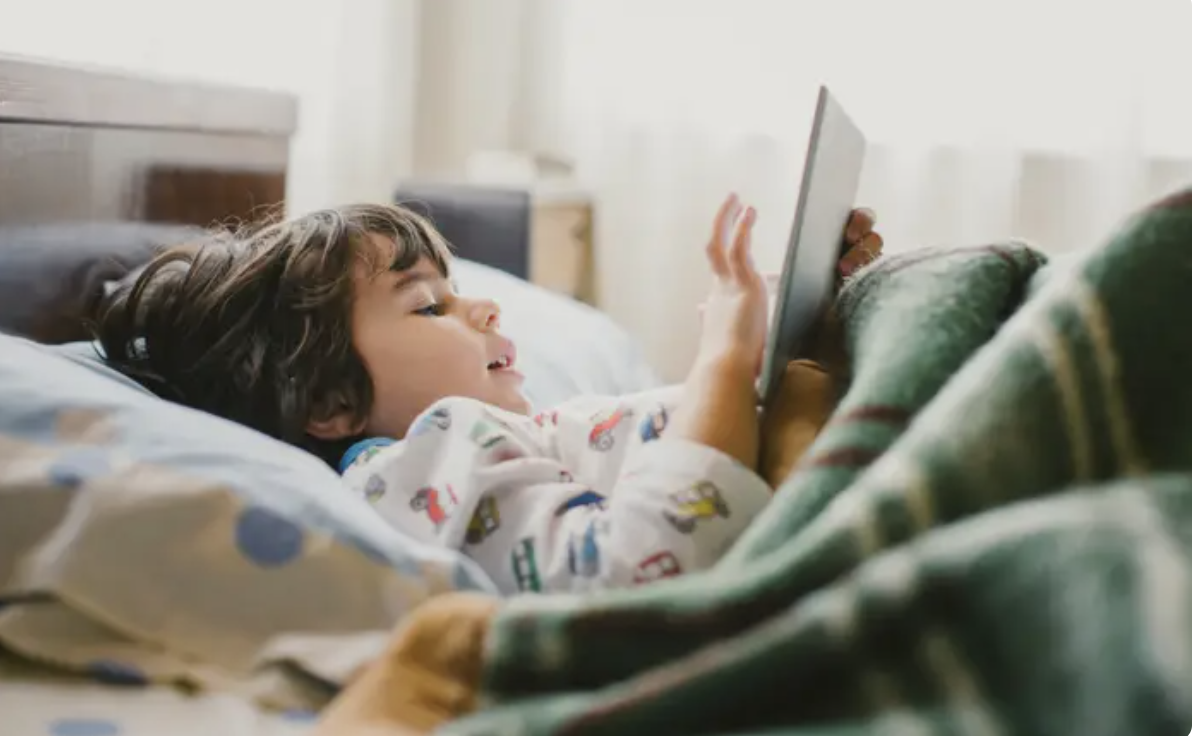 A child is lying in bed under a blanket, smiling while looking at a tablet in their hands