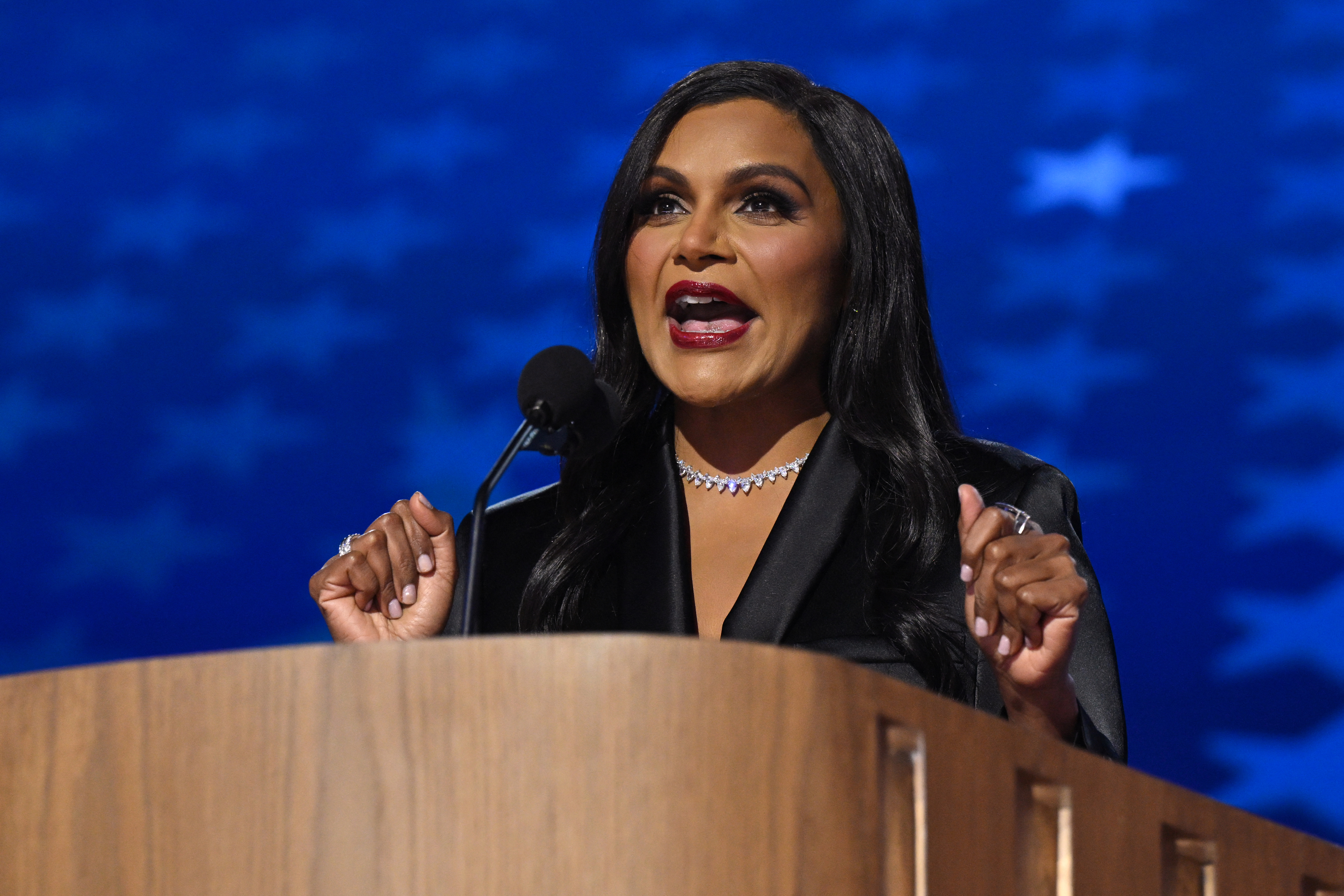 Mindy Kaling speaks passionately at a podium, wearing a chic black outfit and a sparkling necklace, with a blue star-patterned background behind her