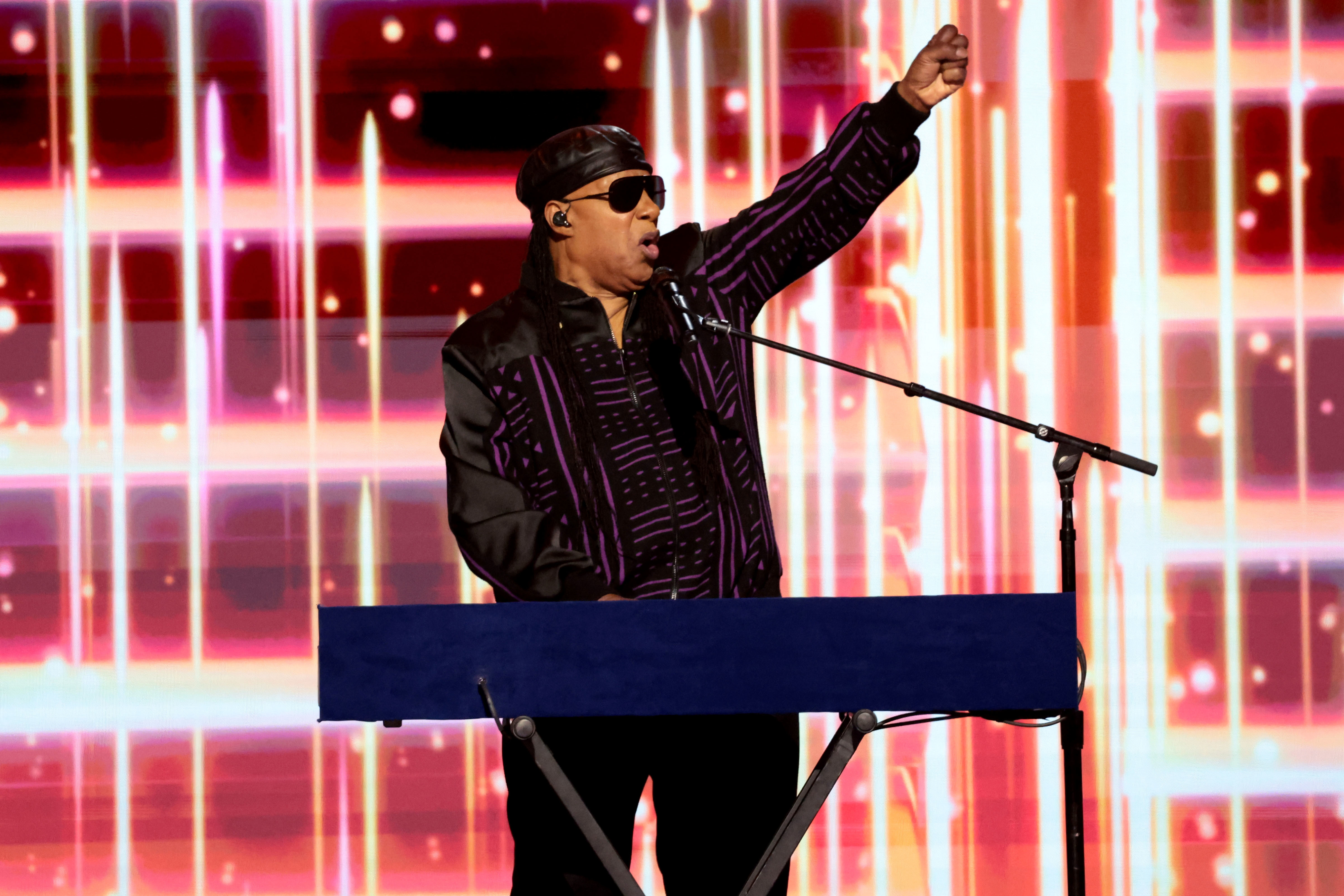 Stevie Wonder performs energetically on stage, standing behind a keyboard with one arm raised, wearing a patterned jacket and sunglasses