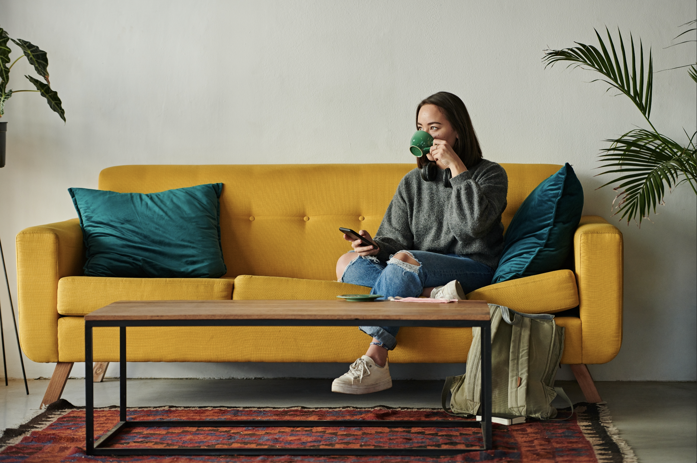 Woman sits on a yellow couch, holding a phone and drinking from a green mug, with a backpack beside her