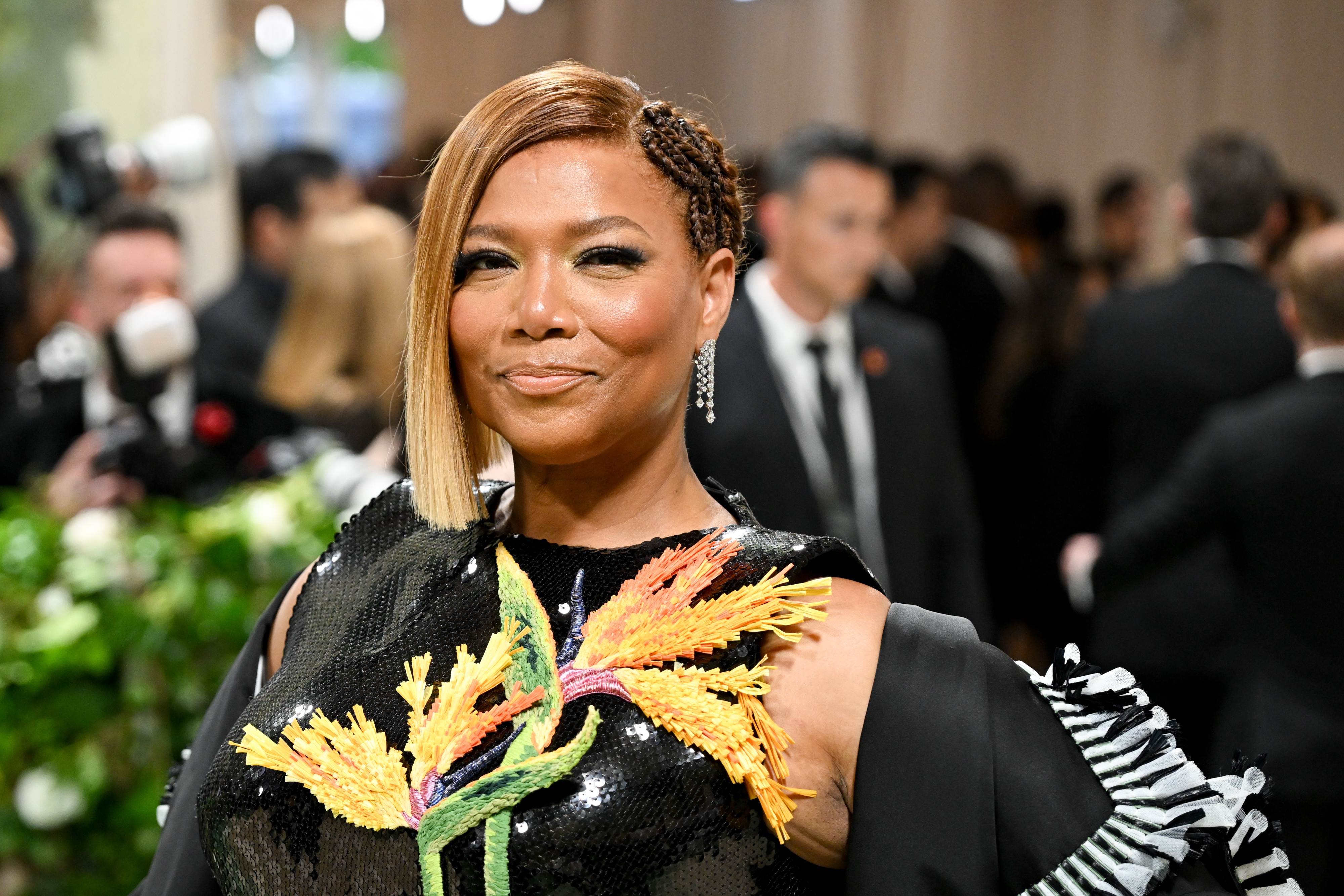 Queen Latifah at an event, wearing a stylish dress adorned with floral designs and black overcoat. Background features a crowd of people