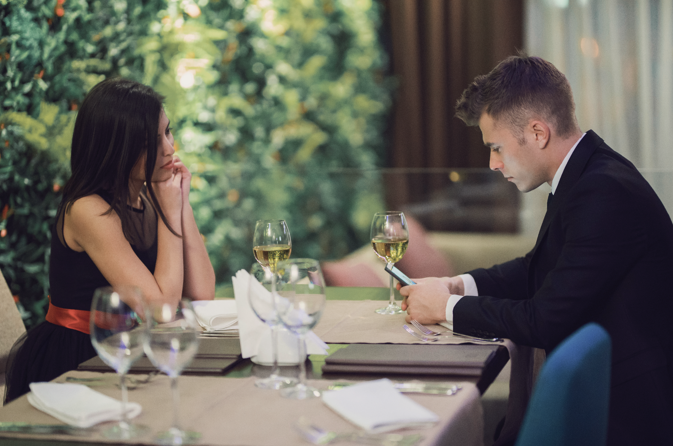 Woman and man dressed formally are at a dining table, she looks at him while he looks at his phone. Two glasses of wine and dining items are on the table
