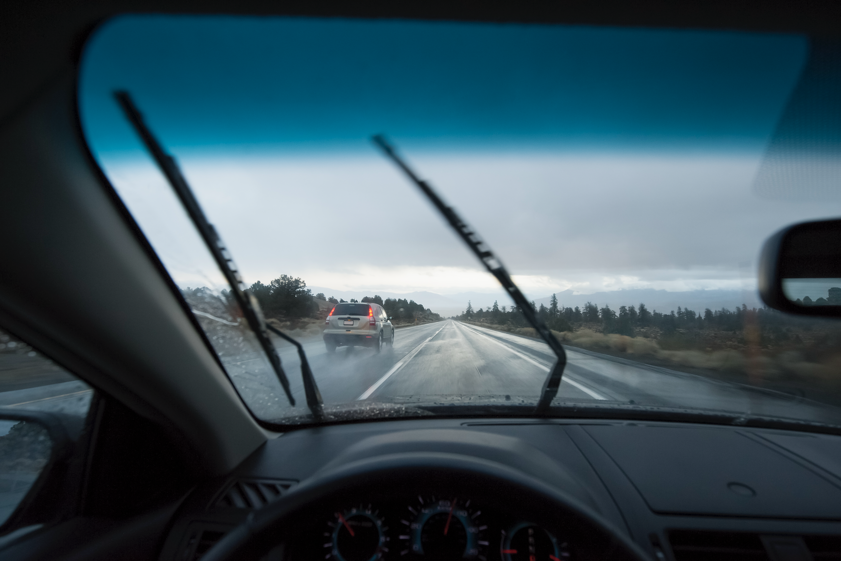 Windshield view of a rainy road, with wipers activated, a car ahead, and a landscape in the distance