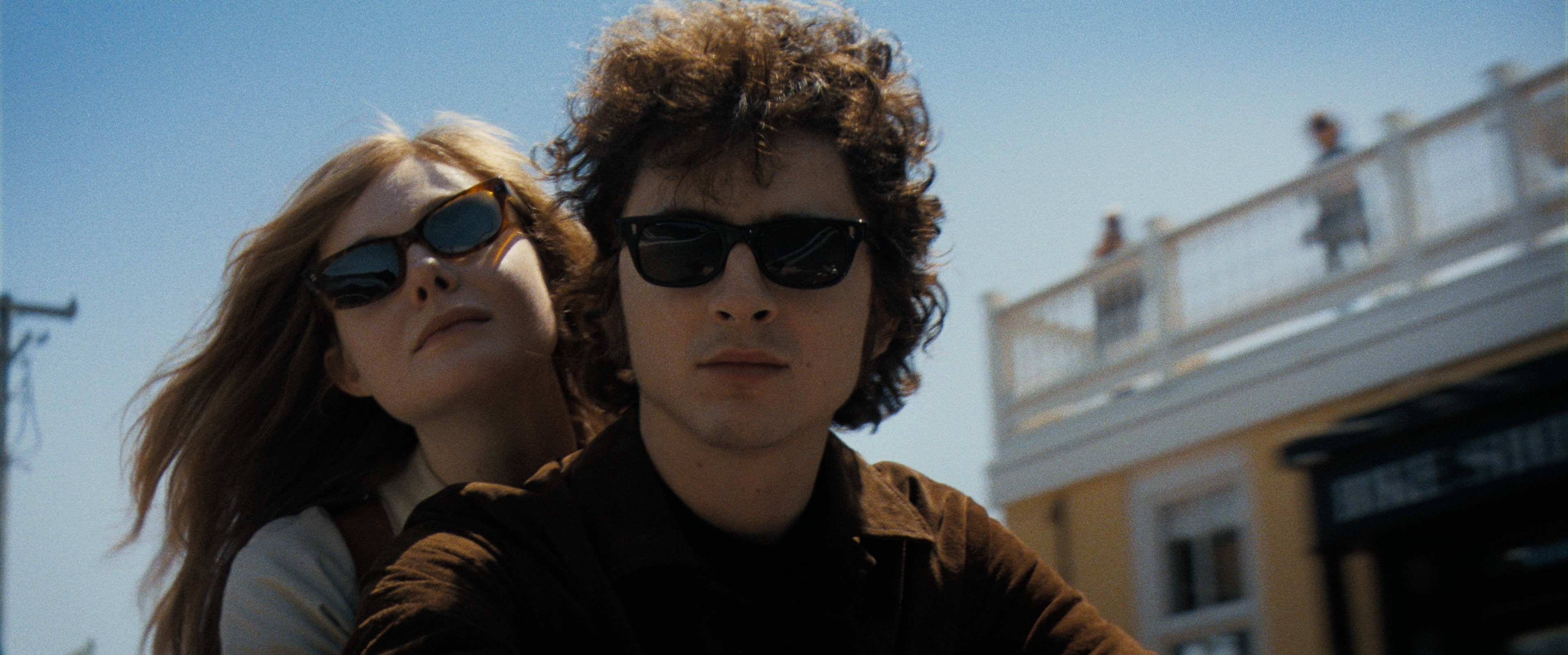 Elle Fanning and Timothée Chalamet ride a scooter; both wear dark sunglasses. The background shows a sunny street with a building and people on a balcony