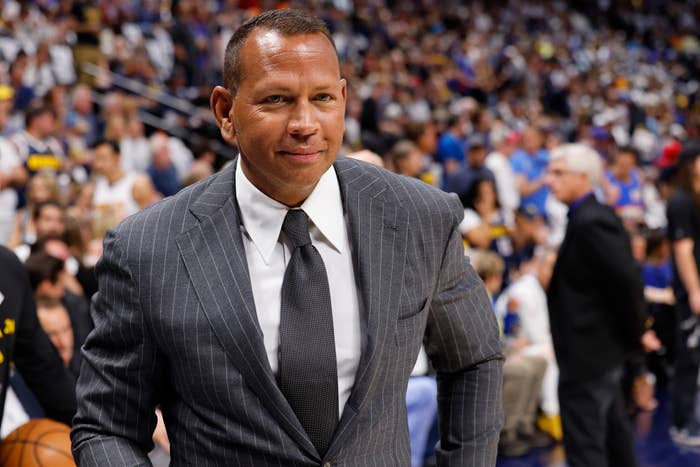 Alex Rodriguez in a pinstripe suit with a tie, attending a sports event with a crowded audience in the background