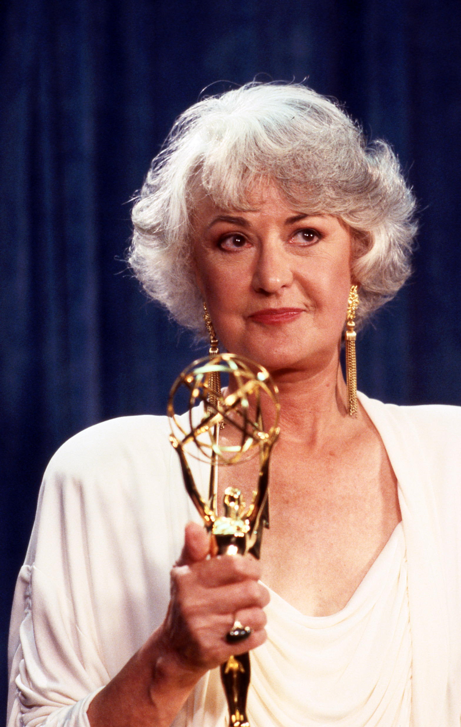 Bea Arthur holds an Emmy award trophy while wearing a draped outfit and long earrings