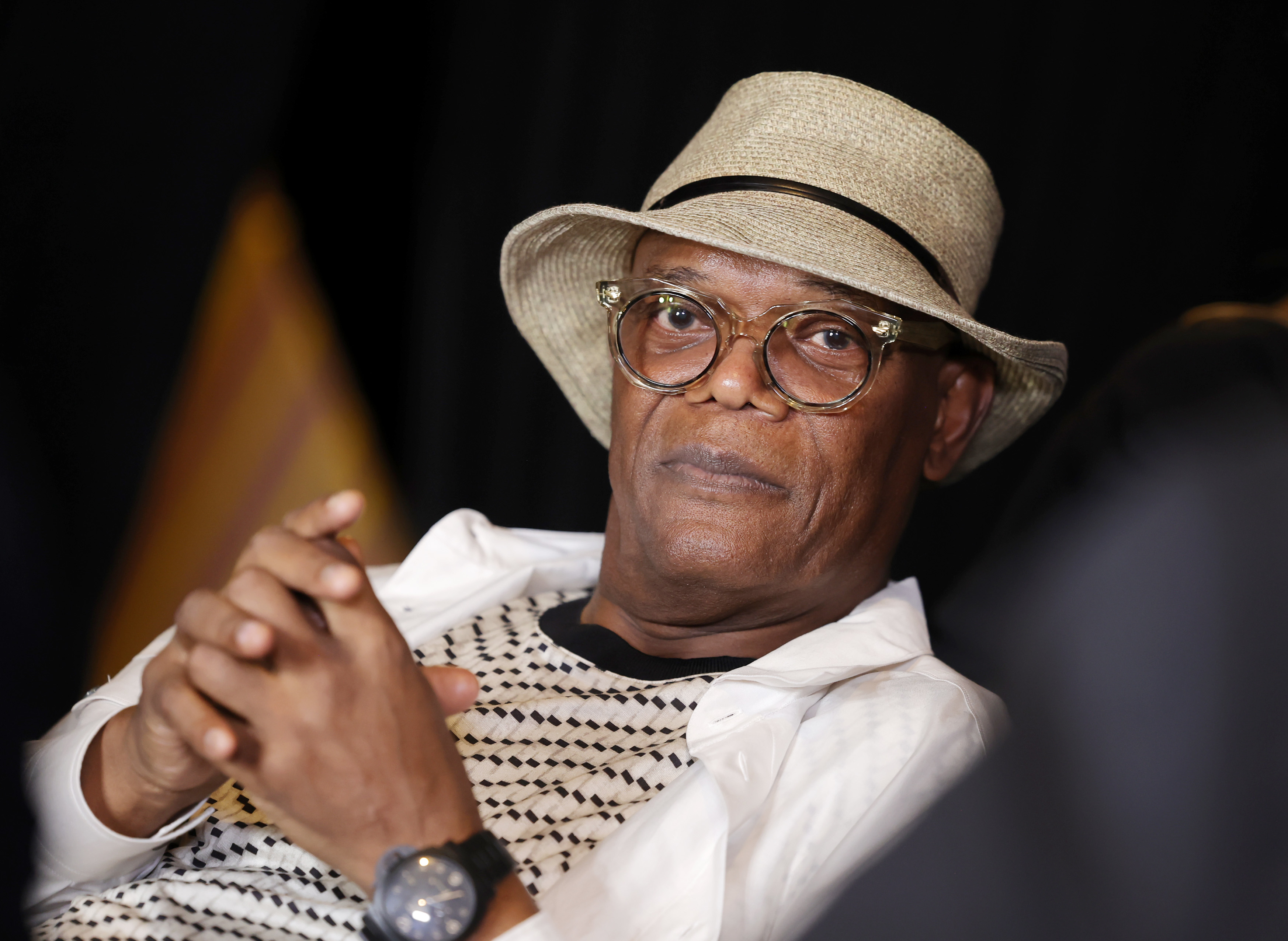 Samuel L. Jackson wearing a patterned shirt, white jacket, glasses, and a hat, while resting his hands together