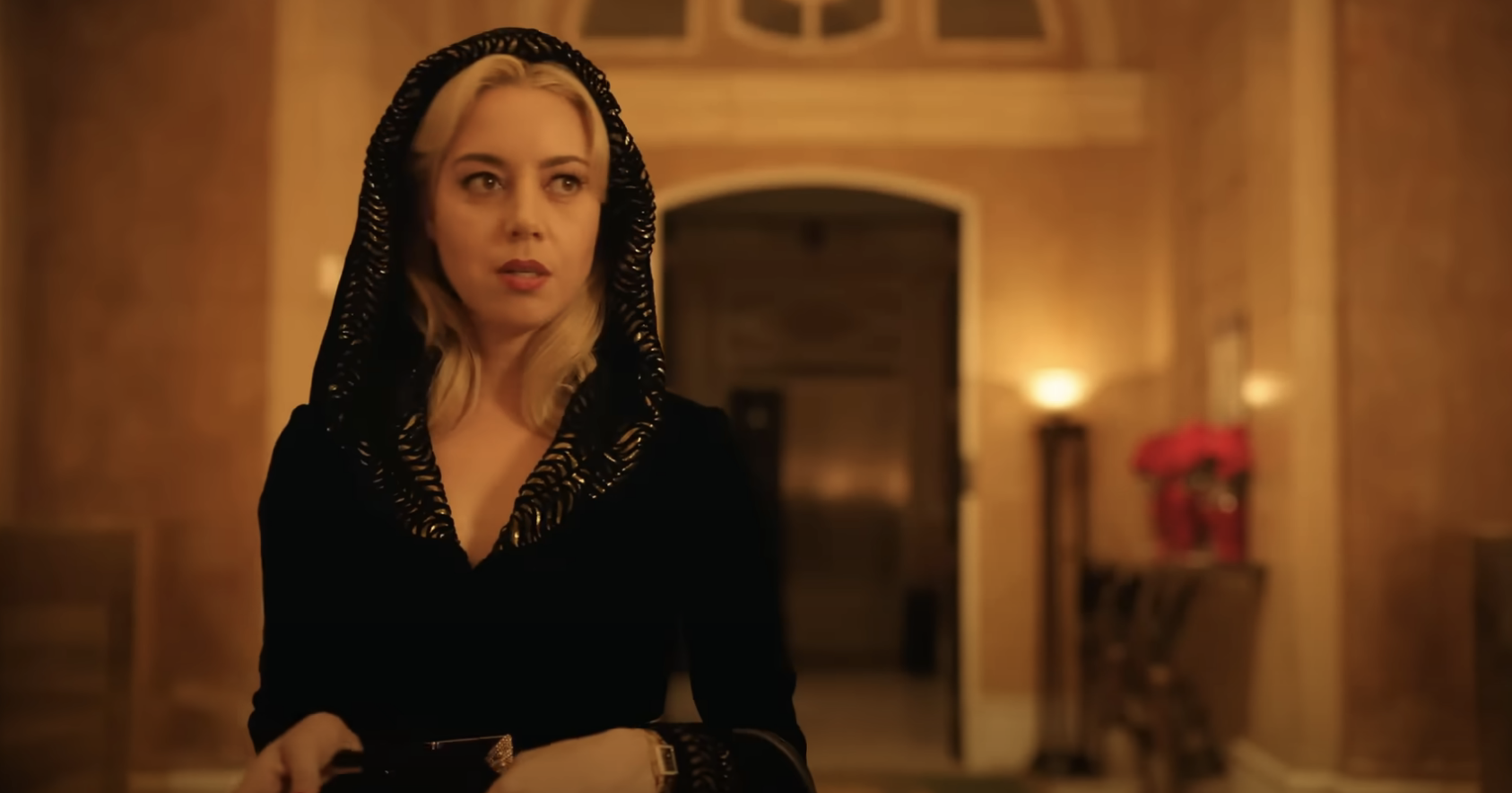 Aubrey Plaza stands indoors, wearing a dark hooded cloak with intricate details, looking to the left. The background is a warmly lit hallway