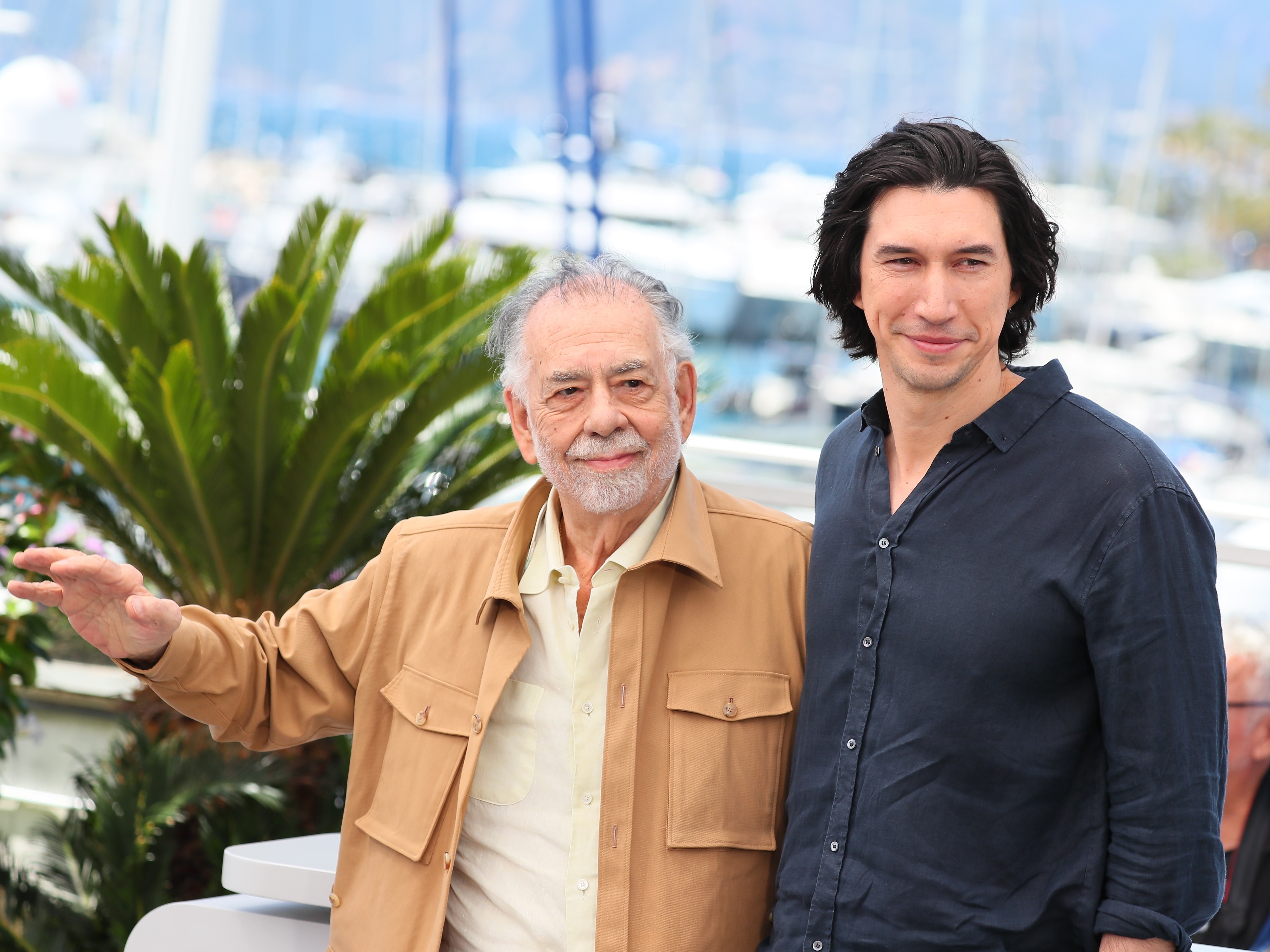 Francis Ford Coppola and Adam Driver pose together outdoors with a marina in the background. Francis wears a beige jacket, and Adam wears a dark shirt