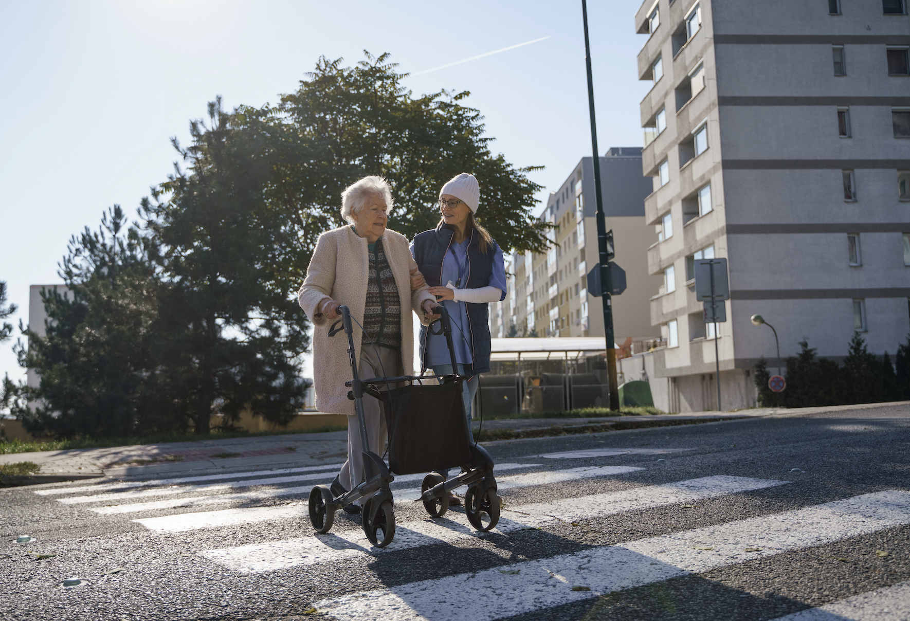 An elderly woman using a walker is helped across a street by a younger woman. Both are smiling and appear to be enjoying the moment
