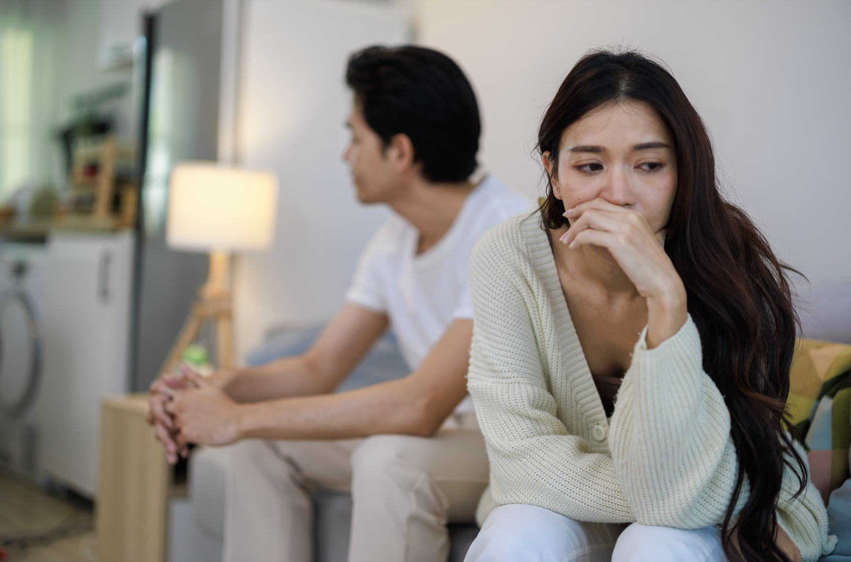A man and a woman sit on a couch, both looking distressed. The man looks away while the woman faces forward with a sad expression. No text present