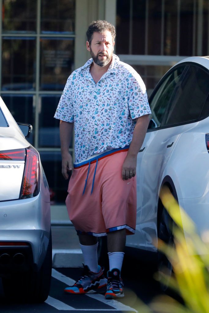 Adam Sandler is seen in casual attire, wearing a patterned shirt, long shorts, socks, and sneakers, walking between parked cars