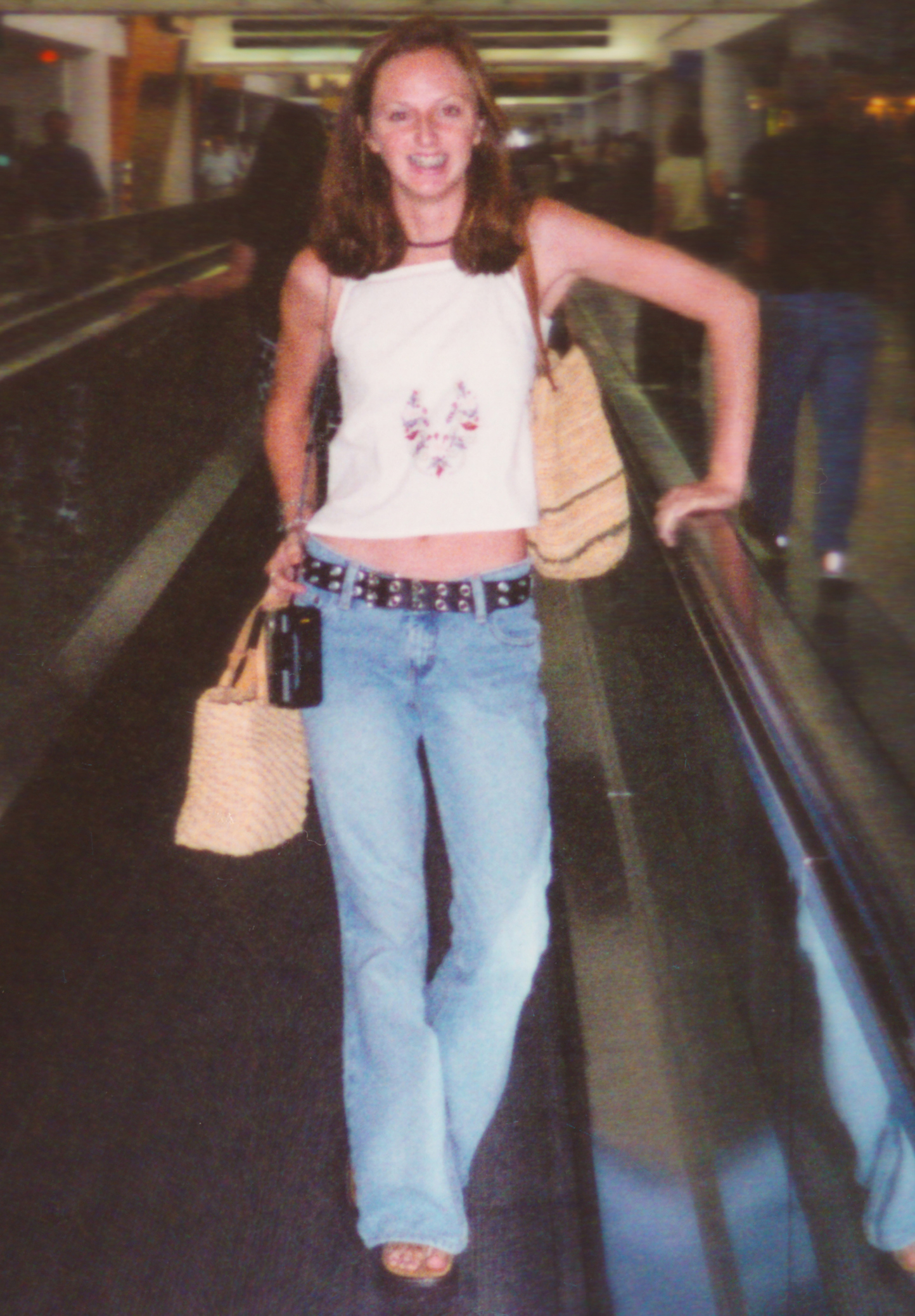 A woman stands on an airport moving walkway, smiling. She is wearing a sleeveless top, blue jeans, and wedge sandals, carrying a woven handbag