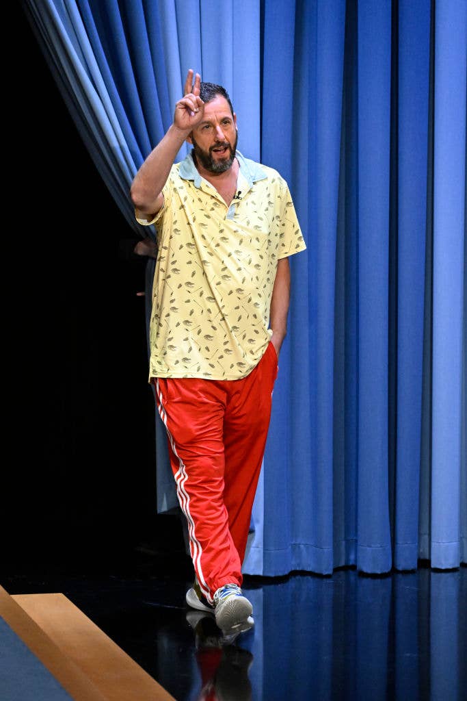 Adam Sandler walking onto a stage, wearing a patterned short-sleeve shirt, red track pants, and sneakers, making a peace sign with his right hand