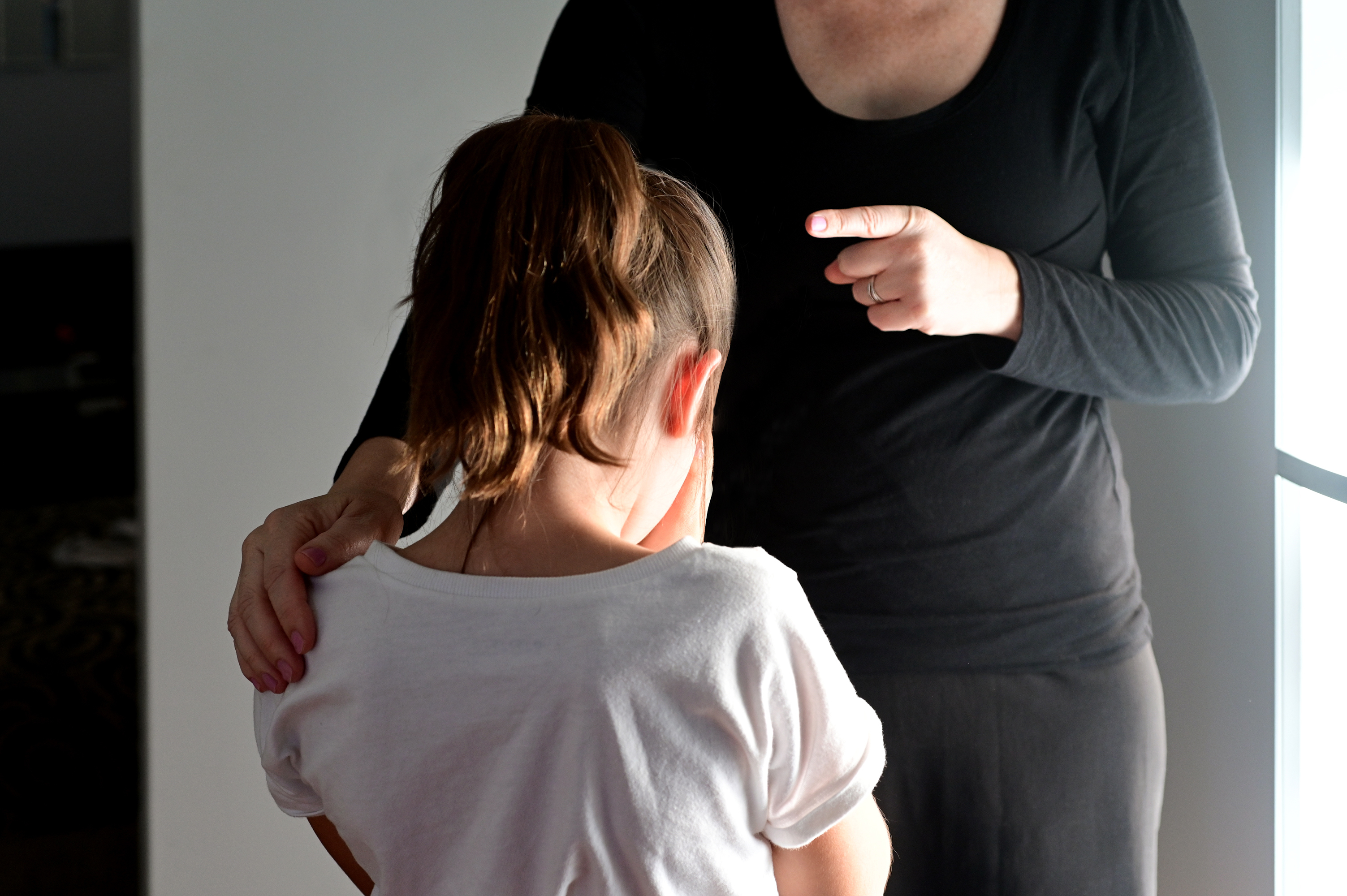 An adult woman points at a young girl standing with her head down and hands covering her face in apparent distress