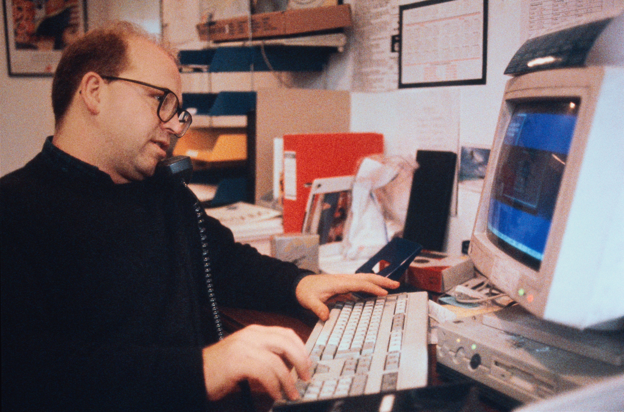 Person using a computer at a desk, with a telephone held to their ear. The desk is cluttered with various office supplies and documents