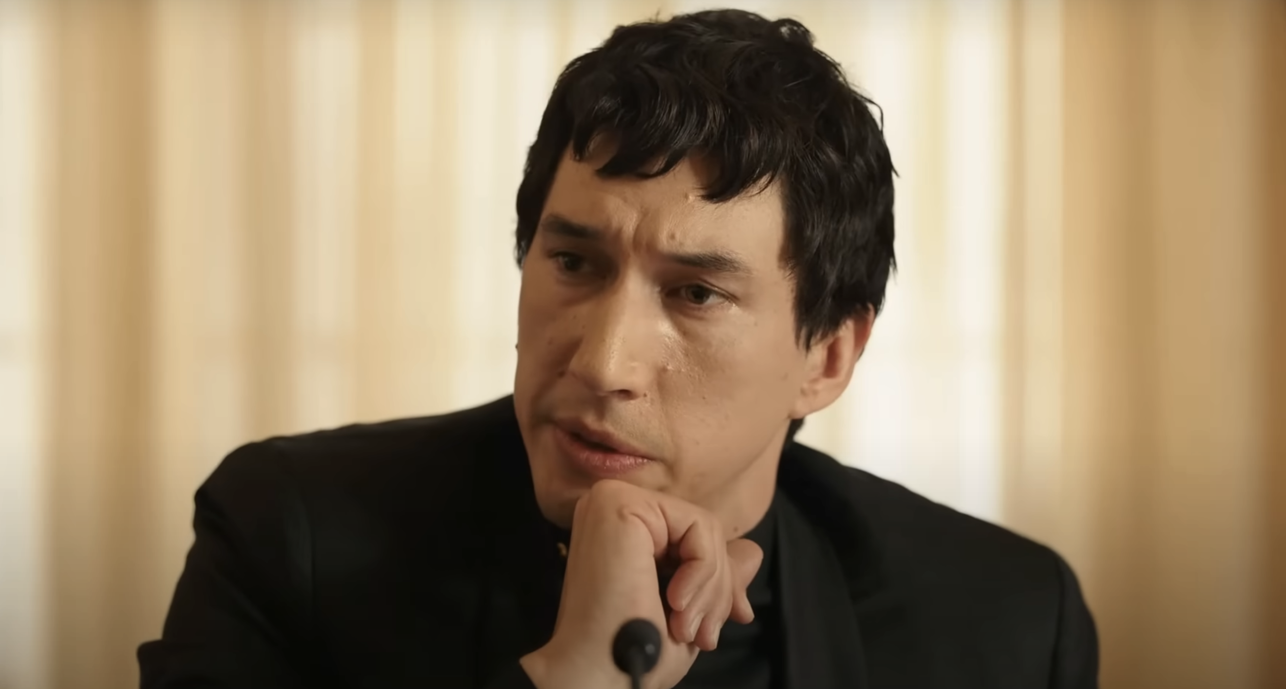 Adam Driver in a scene from a movie or TV show, dressed in a dark suit, resting his chin on his hand and staring thoughtfully into the distance
