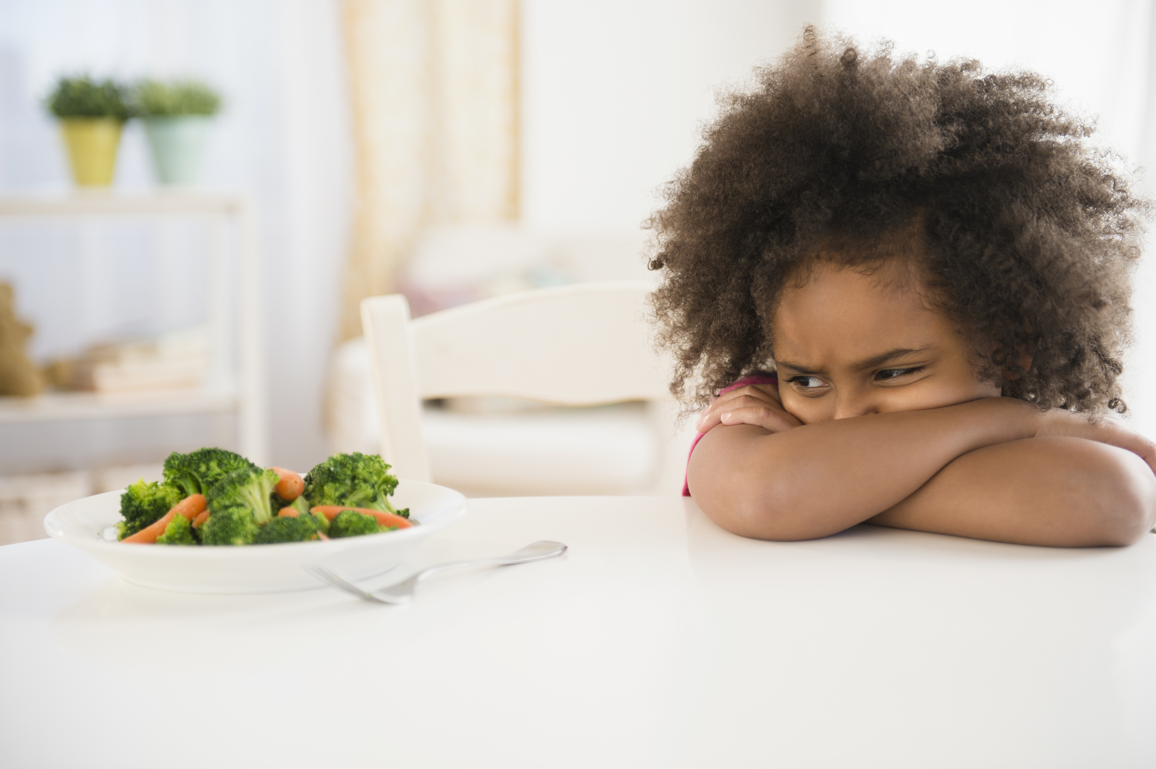 A young child with curly hair looks unhappy at a plate of broccoli and carrots on a white table