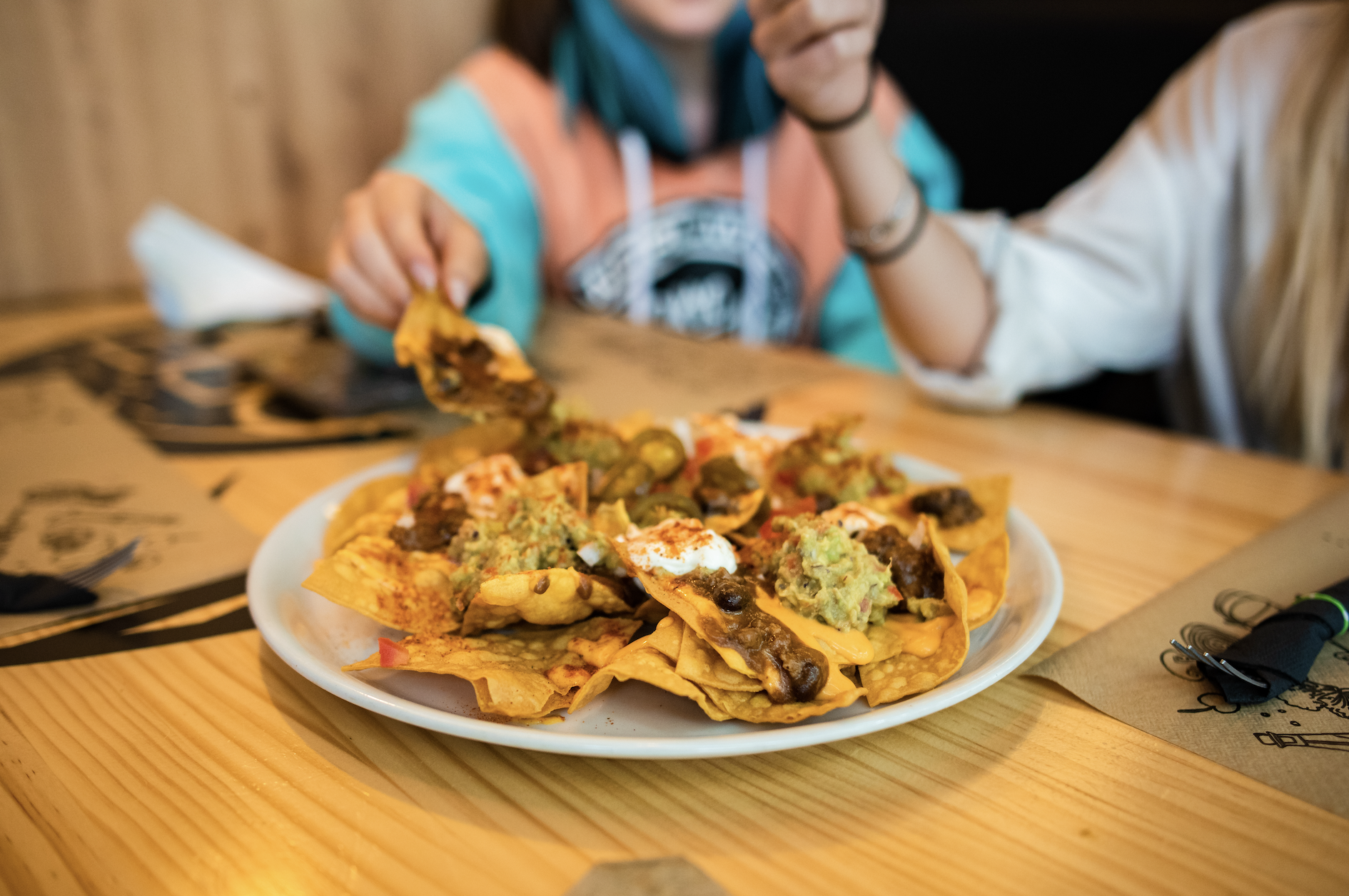 Two unidentified people enjoy a plate of loaded nachos at a wooden table. One is reaching for a nacho, and the other holds a chip near the plate