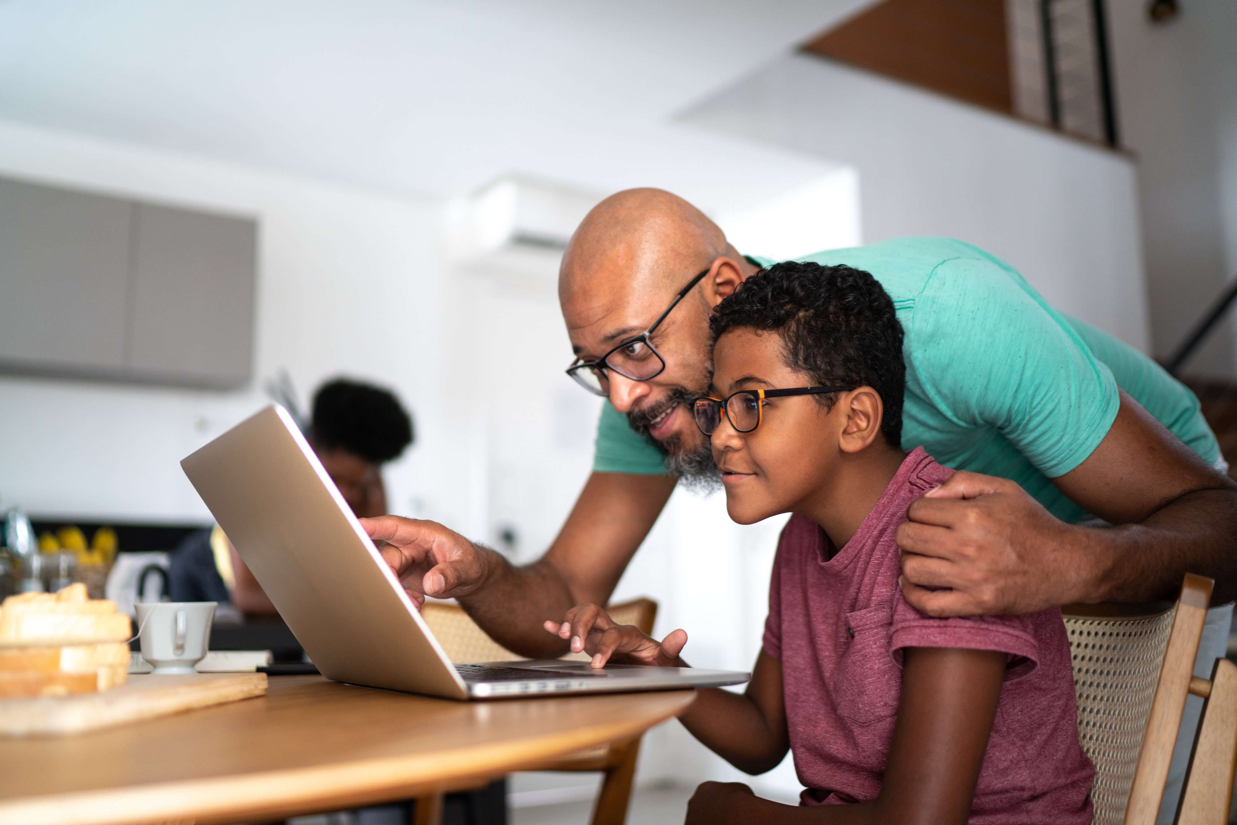 A man and a child, both wearing glasses, smile as they look at a laptop together in a cozy home setting. They exhibit a warm, engaged interaction
