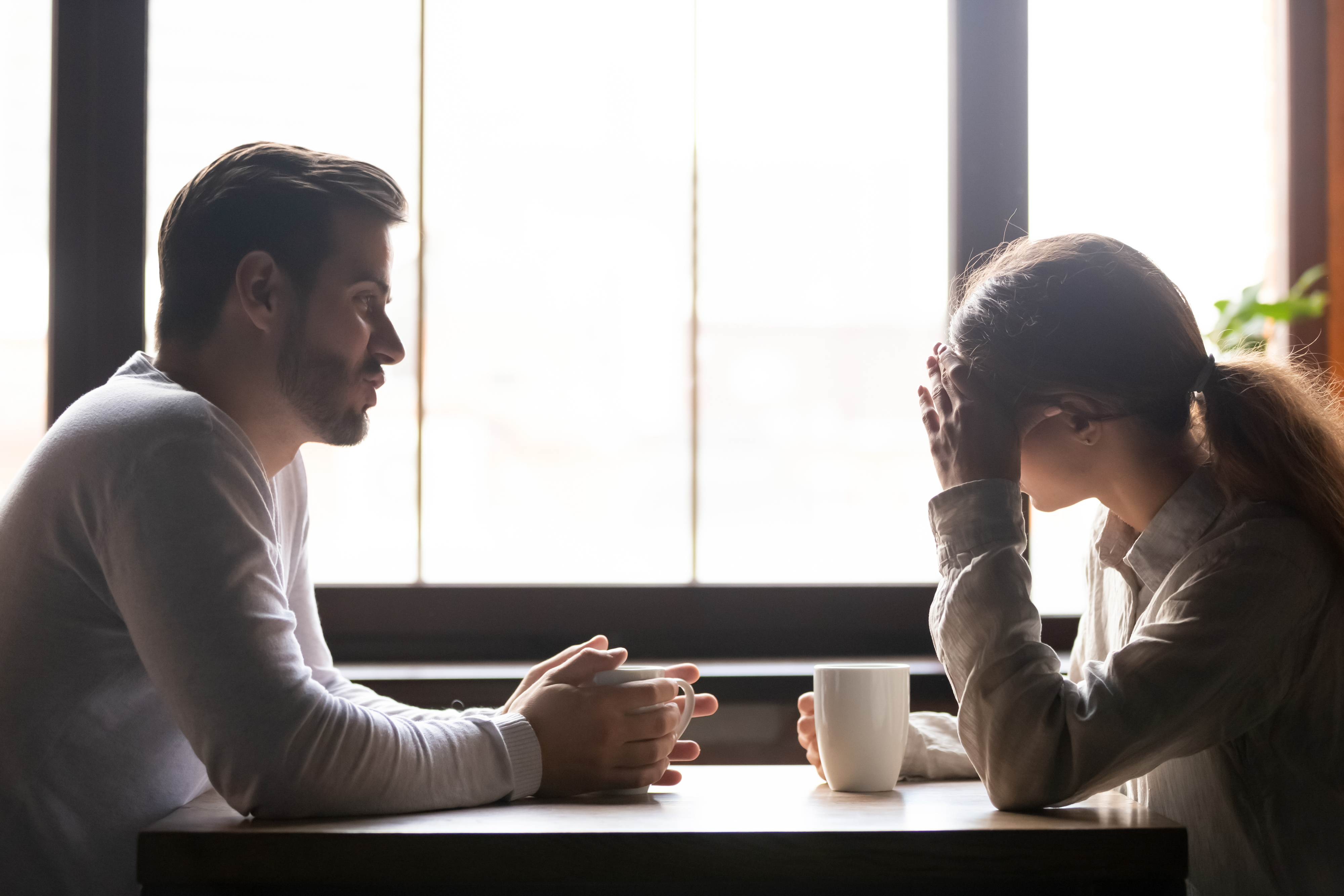 Two people are seated at a table with coffee mugs. The person on the left is looking at the person on the right, who has their head in their hand