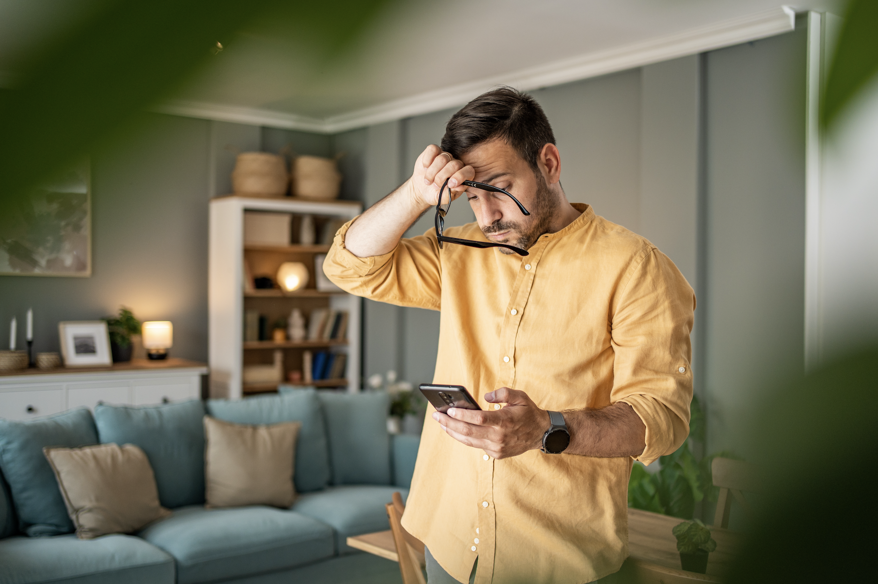 A man stands in his living room, holding a phone in one hand and his glasses in the other, appearing to be deep in thought or confused
