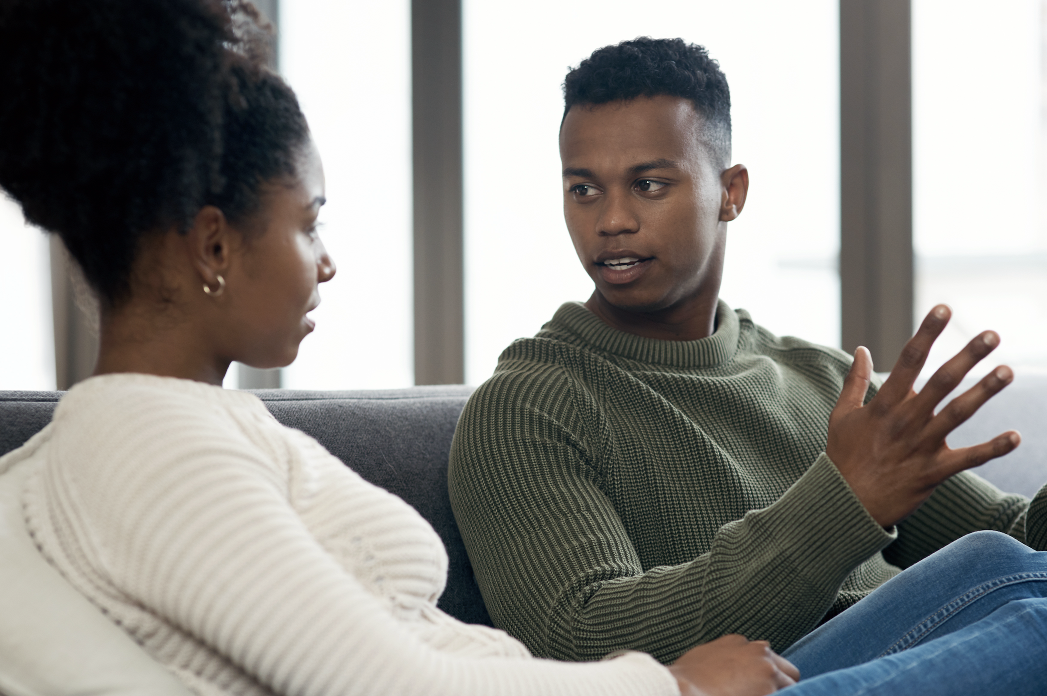 Two people, a woman in a long-sleeve top and a man in a knit sweater, sitting on a couch, engaged in a serious conversation. Names unknown