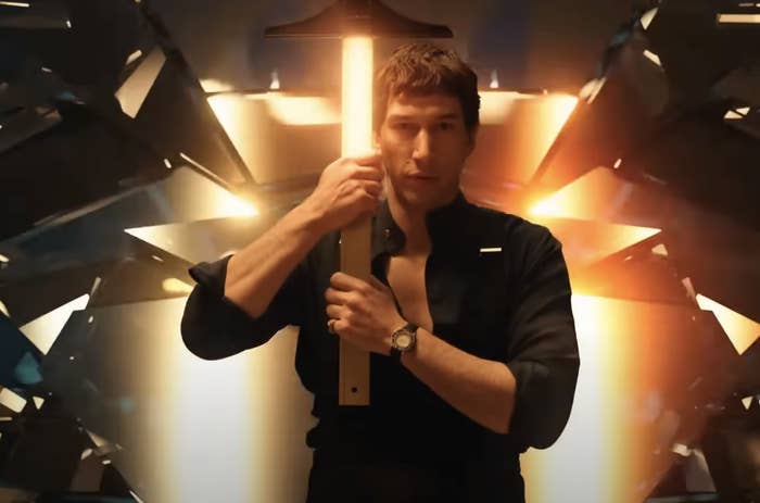 Adam Driver dressed in a dark shirt, holding a glowing, vertical object in a dramatic, metallic set from the movie "Megalopolis"