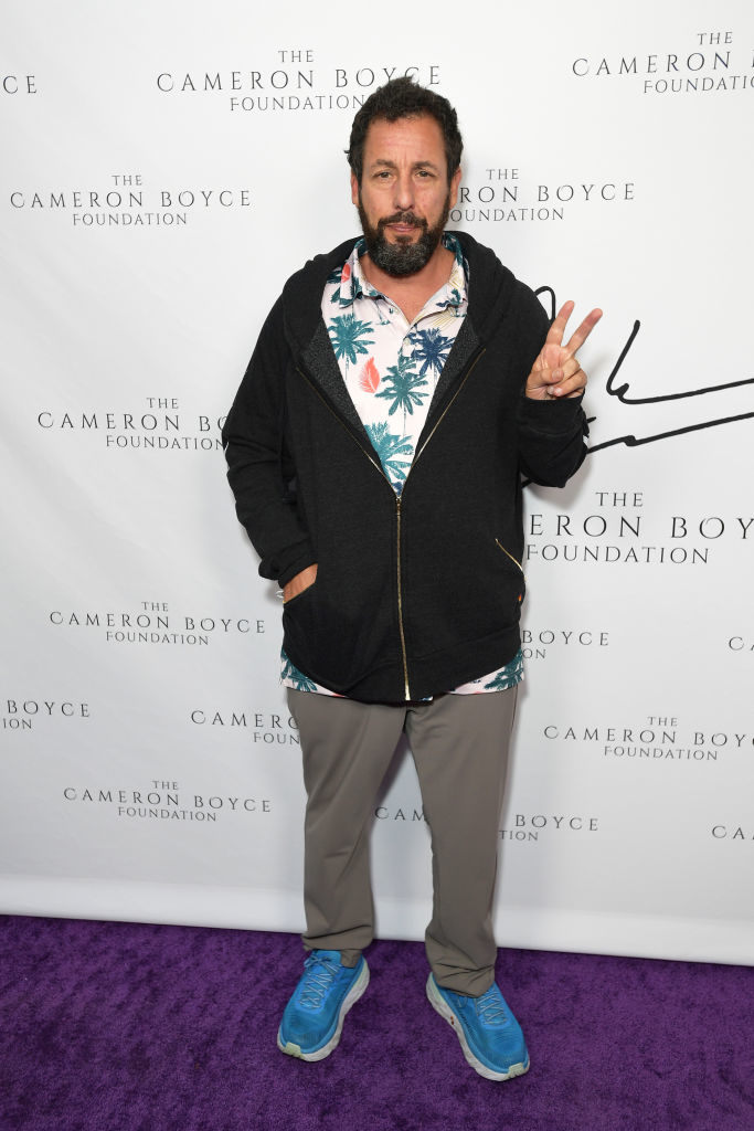 Adam Sandler at The Cameron Boyce Foundation event, wearing a printed shirt, zip-up hoodie, pants, and sneakers, holding up a peace sign