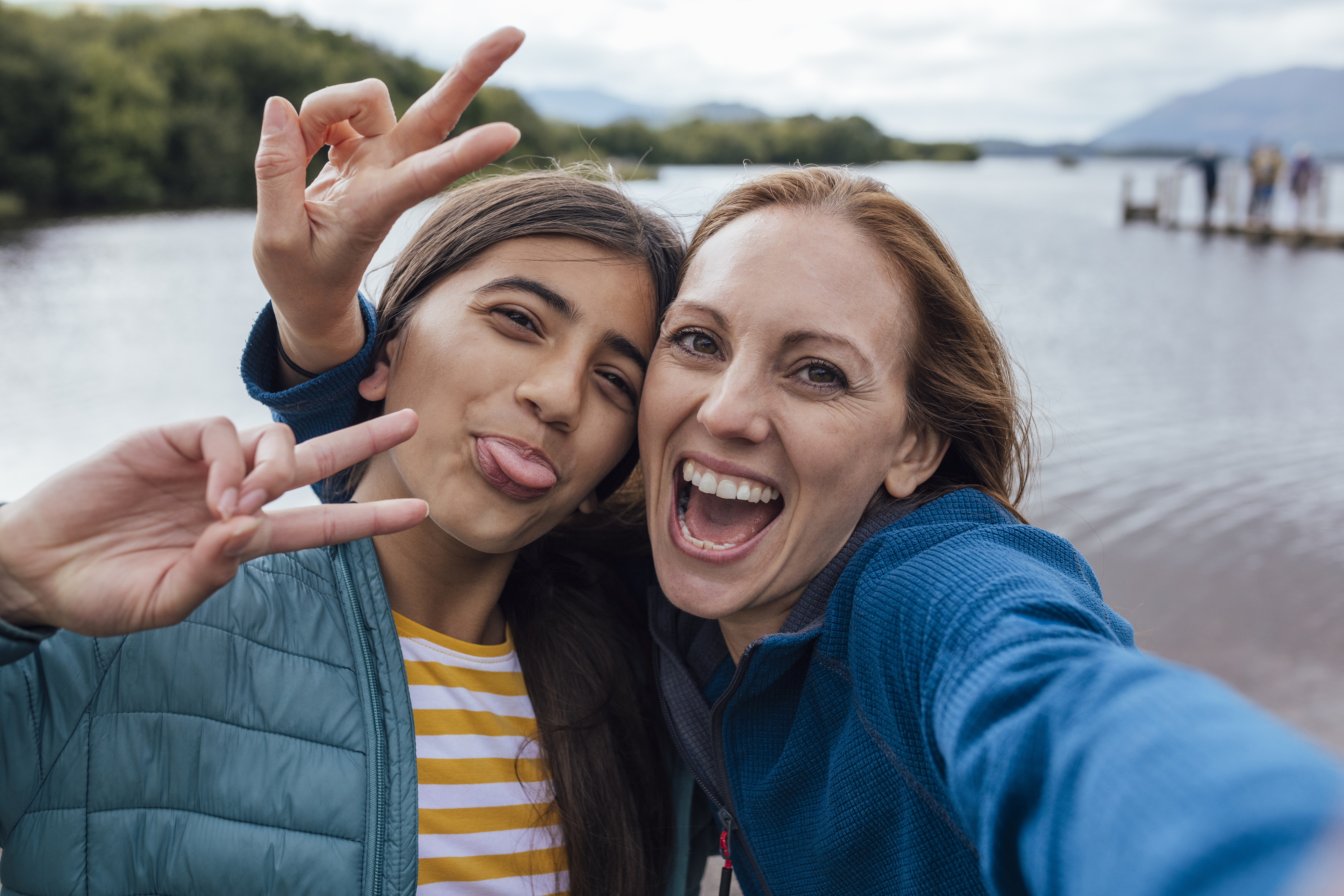 Two people pose for a selfie by a lake. One person sticks out their tongue and makes a peace sign, while the other person smiles widely