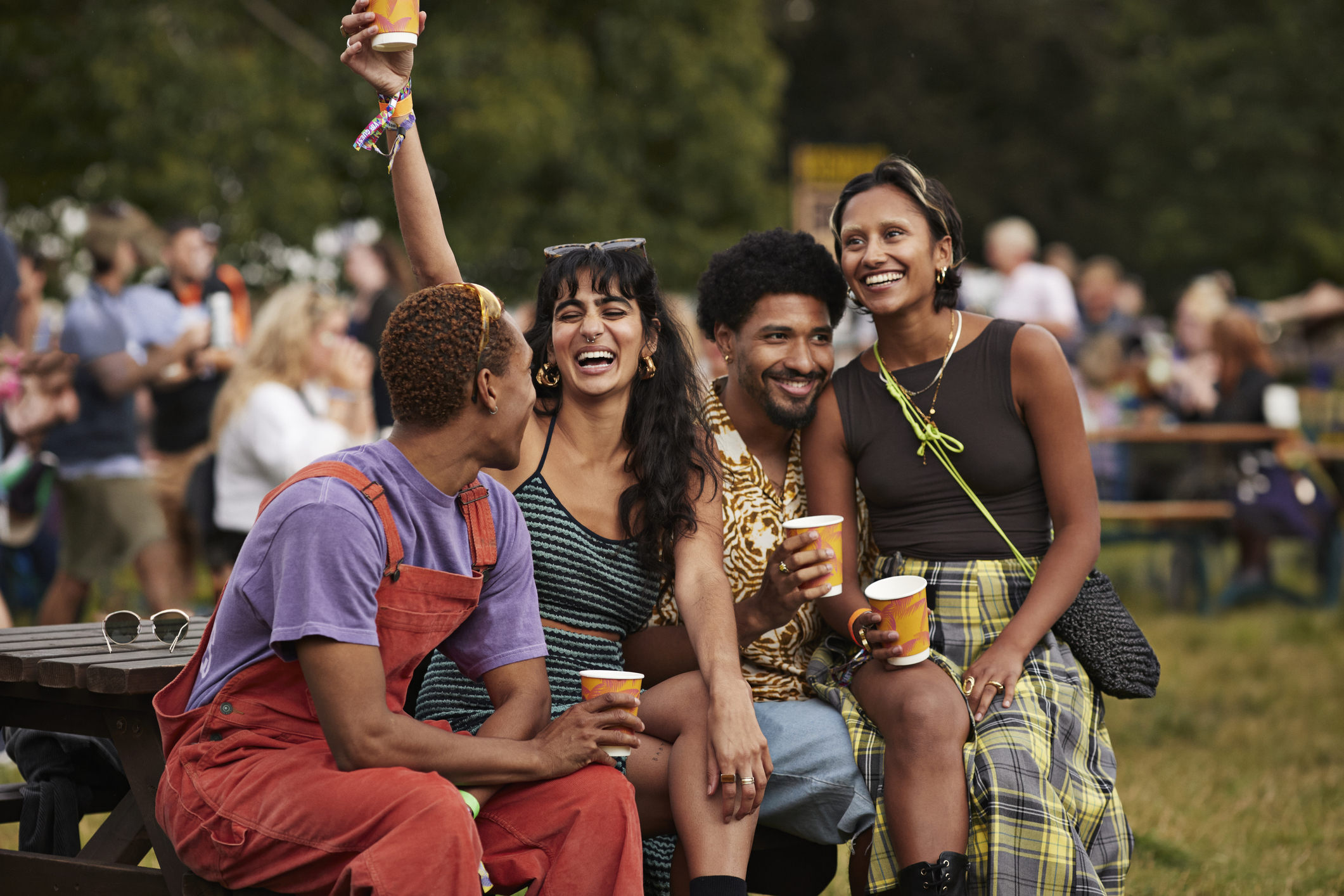 A group of friends enjoy beverages while sitting on a bench at an outdoor gathering, smiling and raising a toast